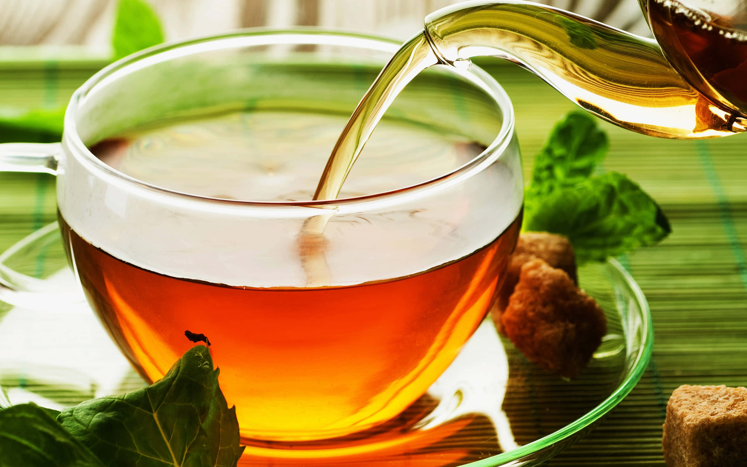 茶 桌布 壁紙 — 金黃色的茶從玻璃壺倒入透明杯中，周圍是新鮮薄荷葉和紅糖塊，放在竹蓆上。