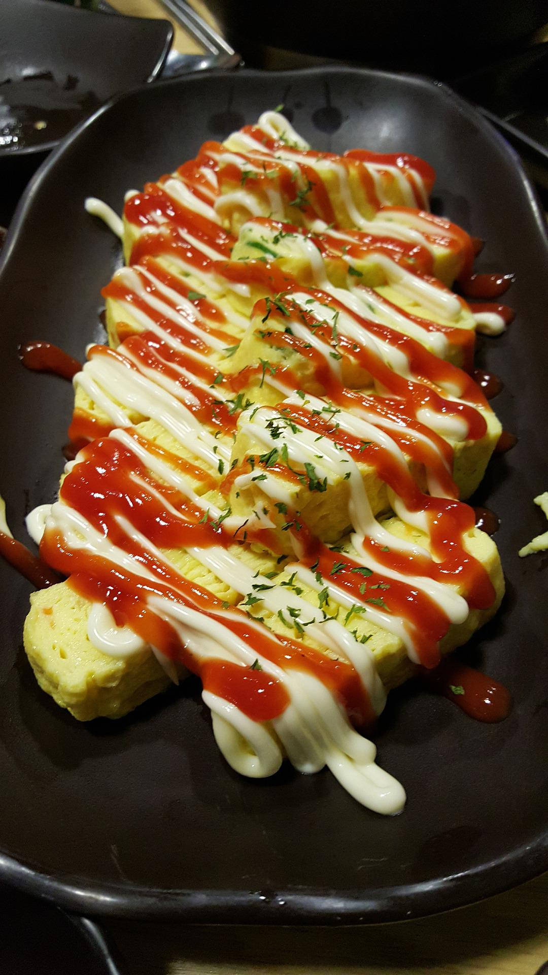 Egg 桌布 壁紙 — 金黃色的蛋捲切片淋上紅色醬汁和白色美乃滋，放在深色盤子上，並以綠色香草點綴。