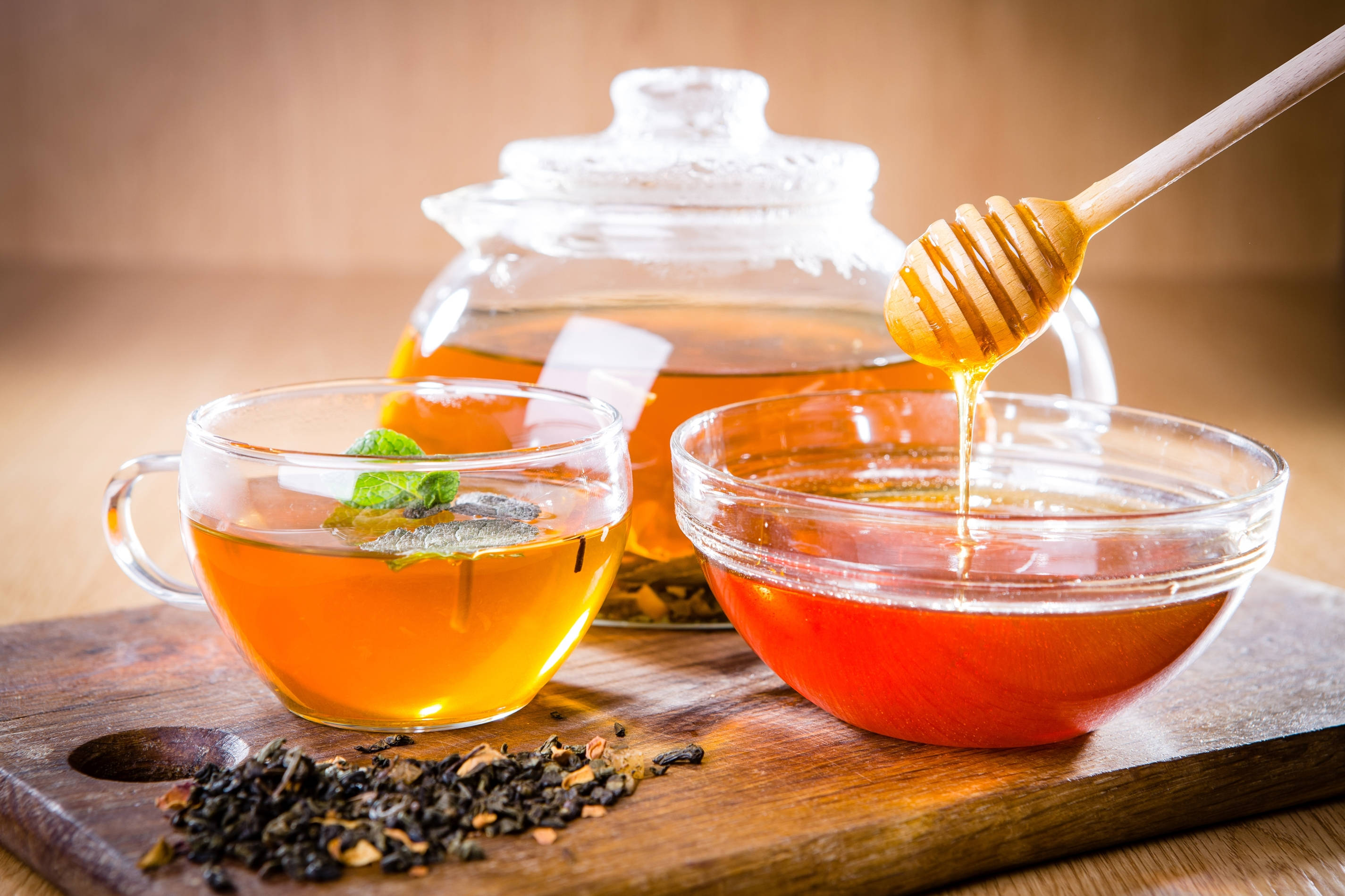 Honeycomb 桌布 壁紙 — 金黃色的蜂蜜從木勺滴入玻璃碗中，旁邊放著一杯琥珀色的茶，配有薄荷和散茶葉，置於質樸的木板上。