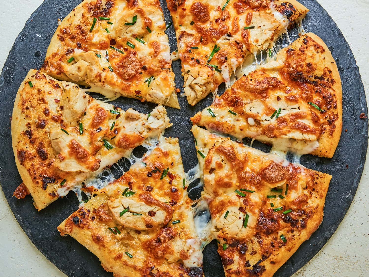 Pizza 桌布 壁紙 — 金黃色的雞肉披薩，配上融化的起司和新鮮的細香蔥，切片放在深色石板服務板上。