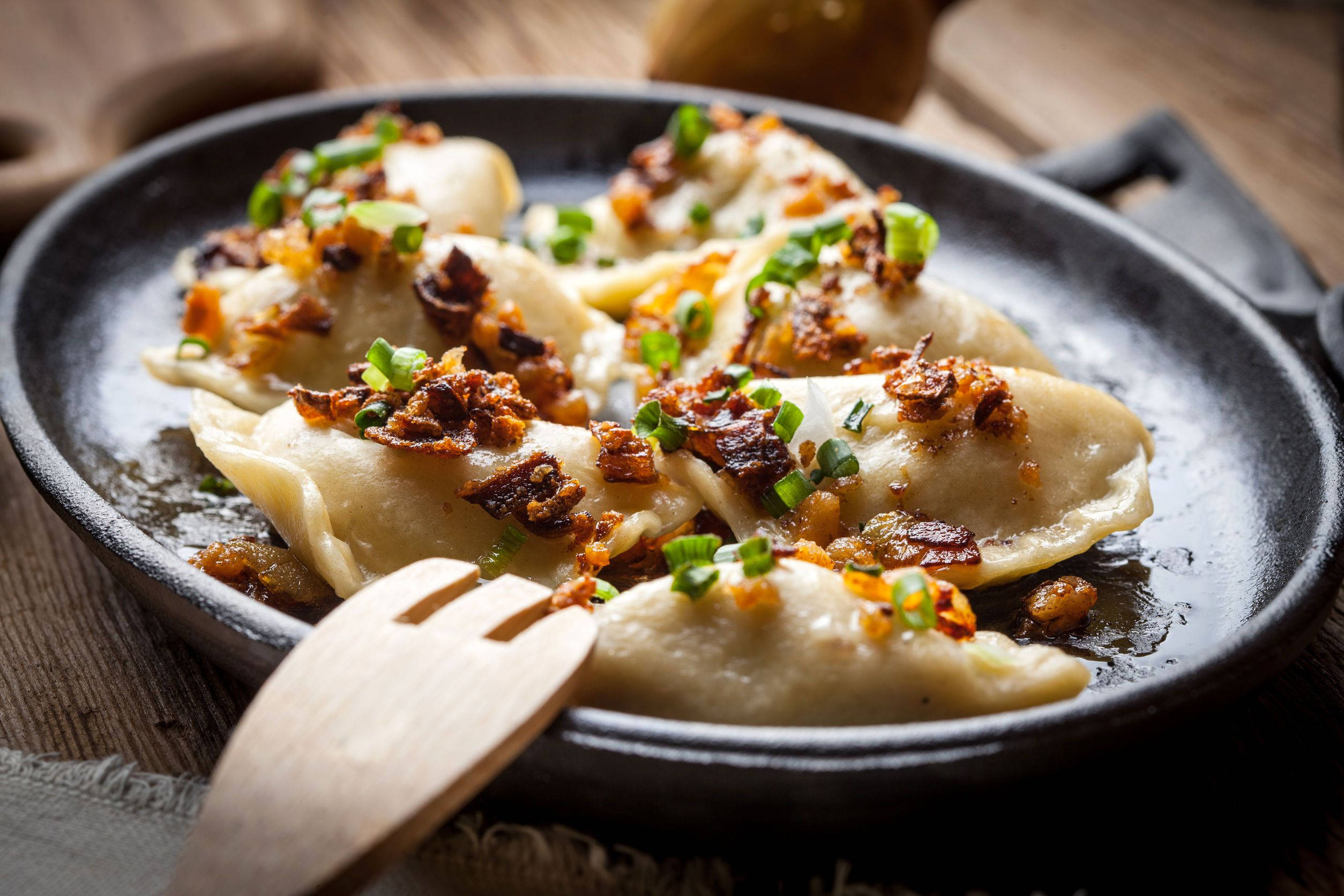 Food Photography 桌布 壁紙 — 金黃色的餃子上面鋪滿酥脆炸洋蔥和蔥花，放在木桌上的黑色煎鍋中。