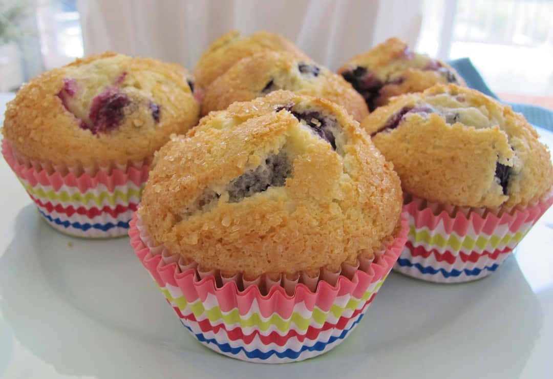 Blueberry Muffins 桌布 壁紙 — 金黃色藍莓馬芬放在色彩繽紛的條紋紙托中，整齊地擺放在明亮的窗邊白色盤子上，令人垂涎。