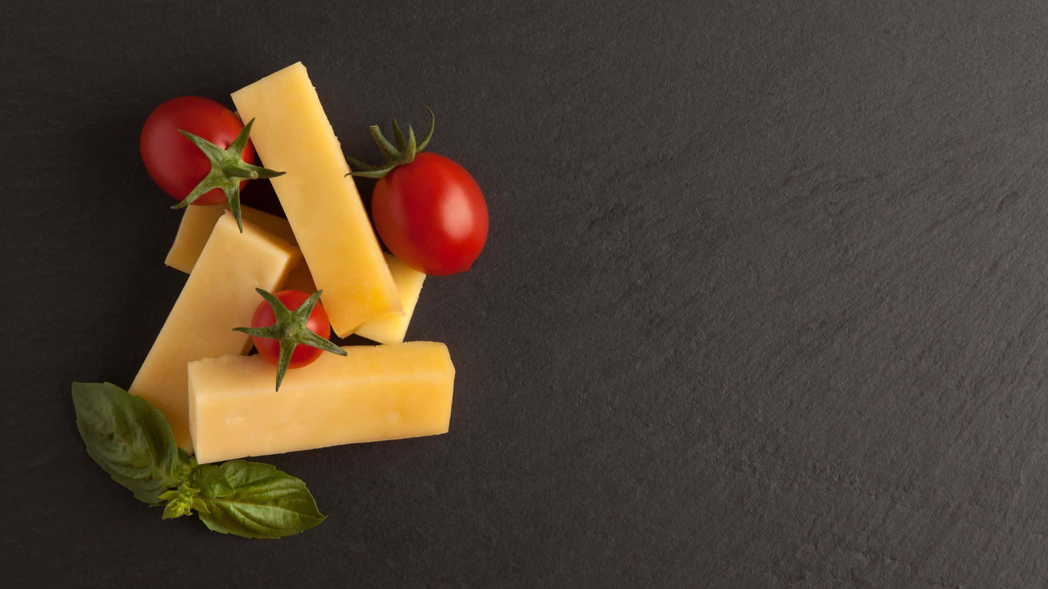 Cheese 桌布 壁紙 — 金黃色起司片和鮮豔的櫻桃番茄配羅勒葉，排列在深色石板表面上。