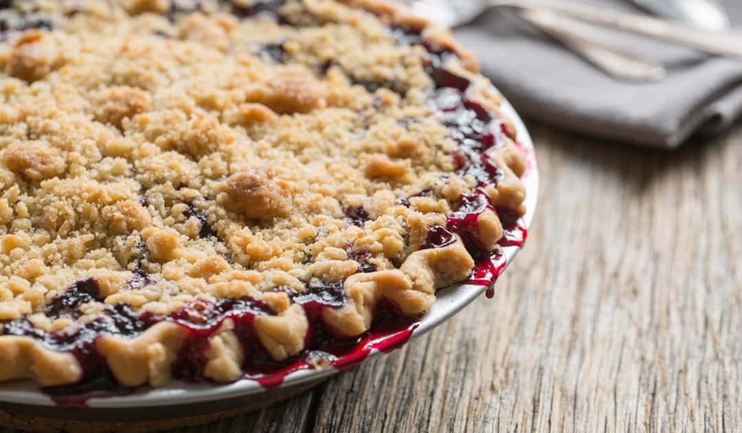 Blueberry Pie 桌布 壁紙 — 金黃色酥脆金寶藍莓派的特寫，果汁滲出在質樸的木桌上。