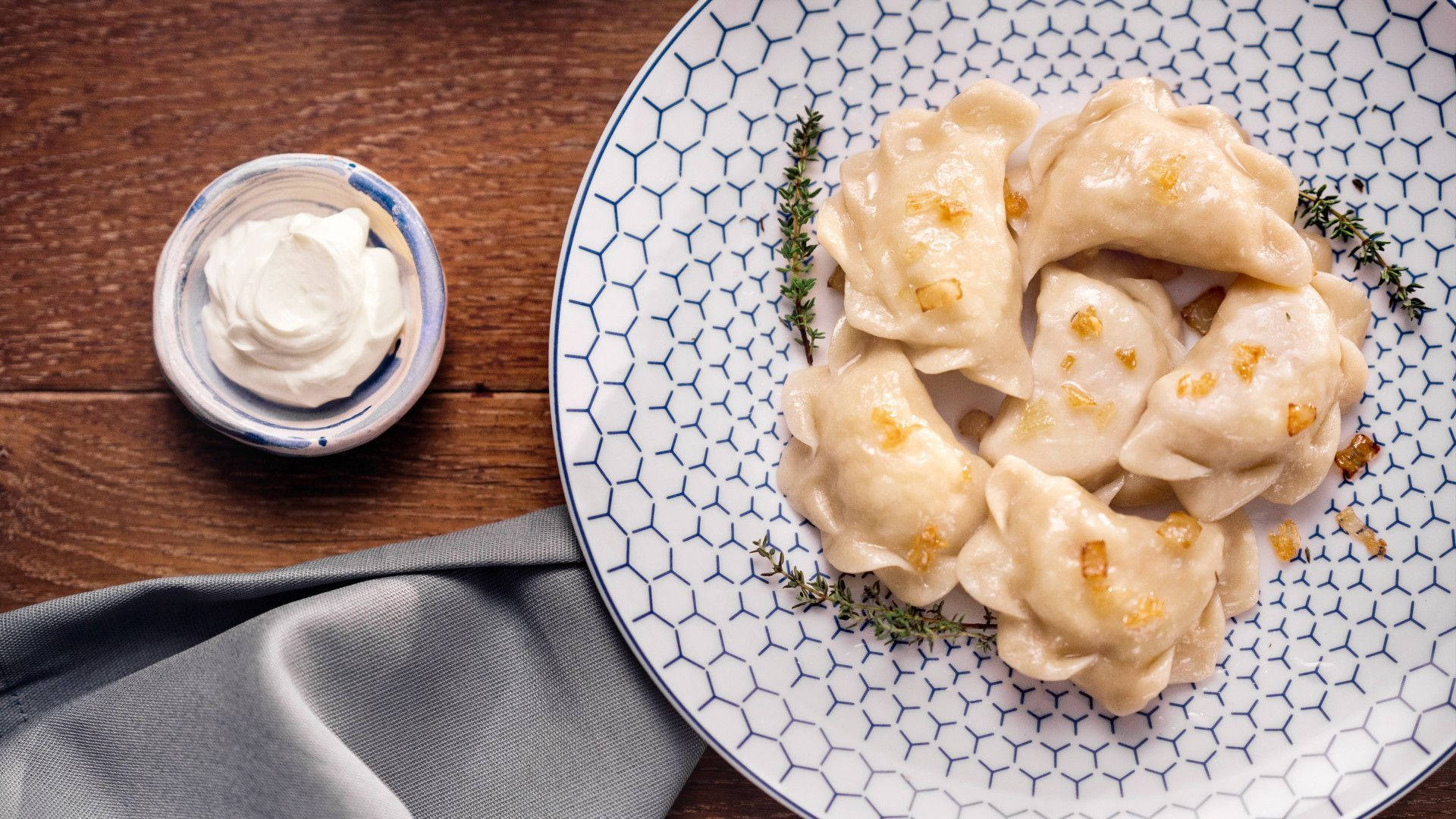 Food Photography 桌布 壁紙 — 金黃色餃子飾以百里香和脆洋蔥，放在藍色六角形盤上，旁邊有一勺酸奶油，置於鄉村風木桌上。