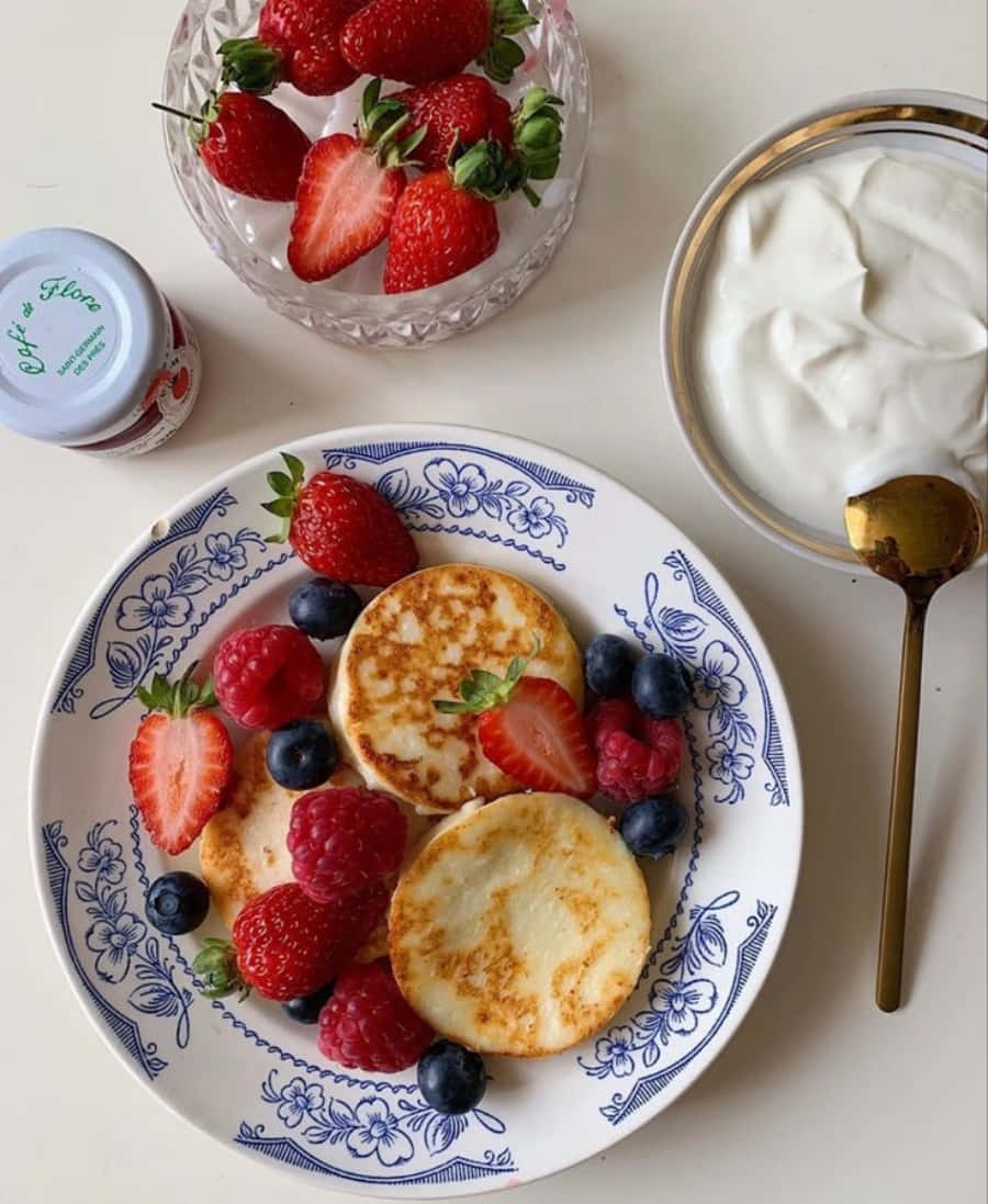 Aesthetic Food 桌布 壁紙 — 金黃色鬆餅上鋪滿新鮮草莓、覆盆子和藍莓，放在藍色花卉圖案盤子上，旁邊有一碗優格和一罐果醬。
