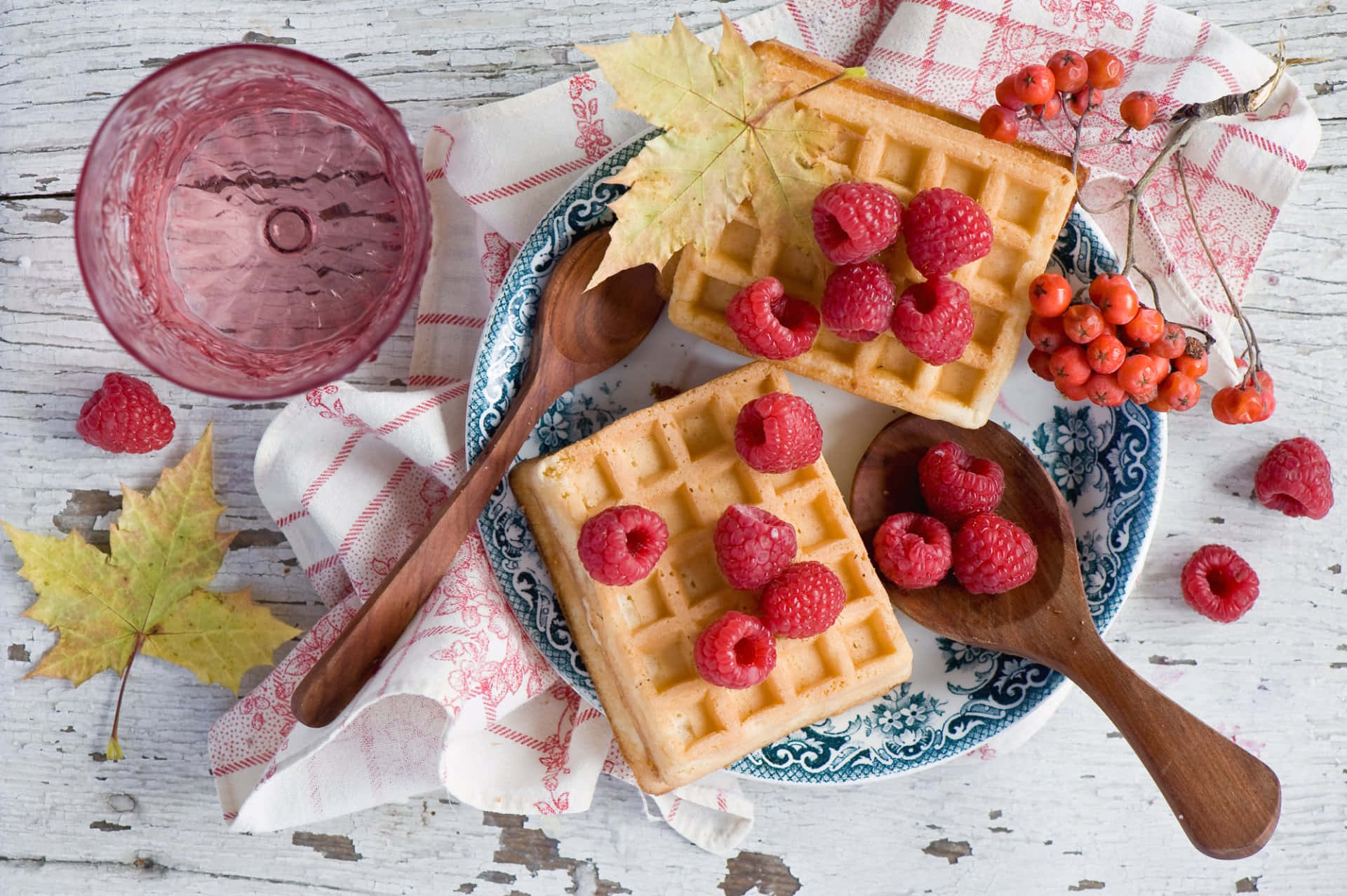 Waffle 桌布 壁紙 — 金黃色鬆餅搭配新鮮覆盆子和秋葉，置於花紋盤上，旁邊有粉紅色玻璃杯和木匙。
