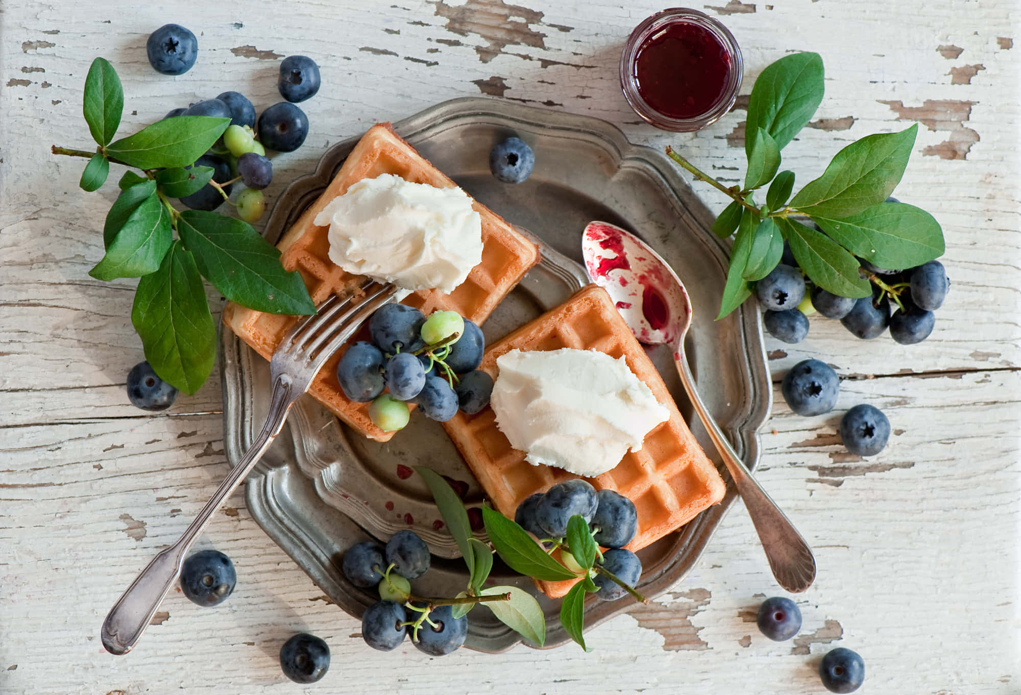 Waffle 桌布 壁紙 — 金黃色鬆餅搭配鮮奶油和新鮮藍莓，放置在一個暗淡的銀托盤上，周圍環繞著綠葉和一罐莓果醬。