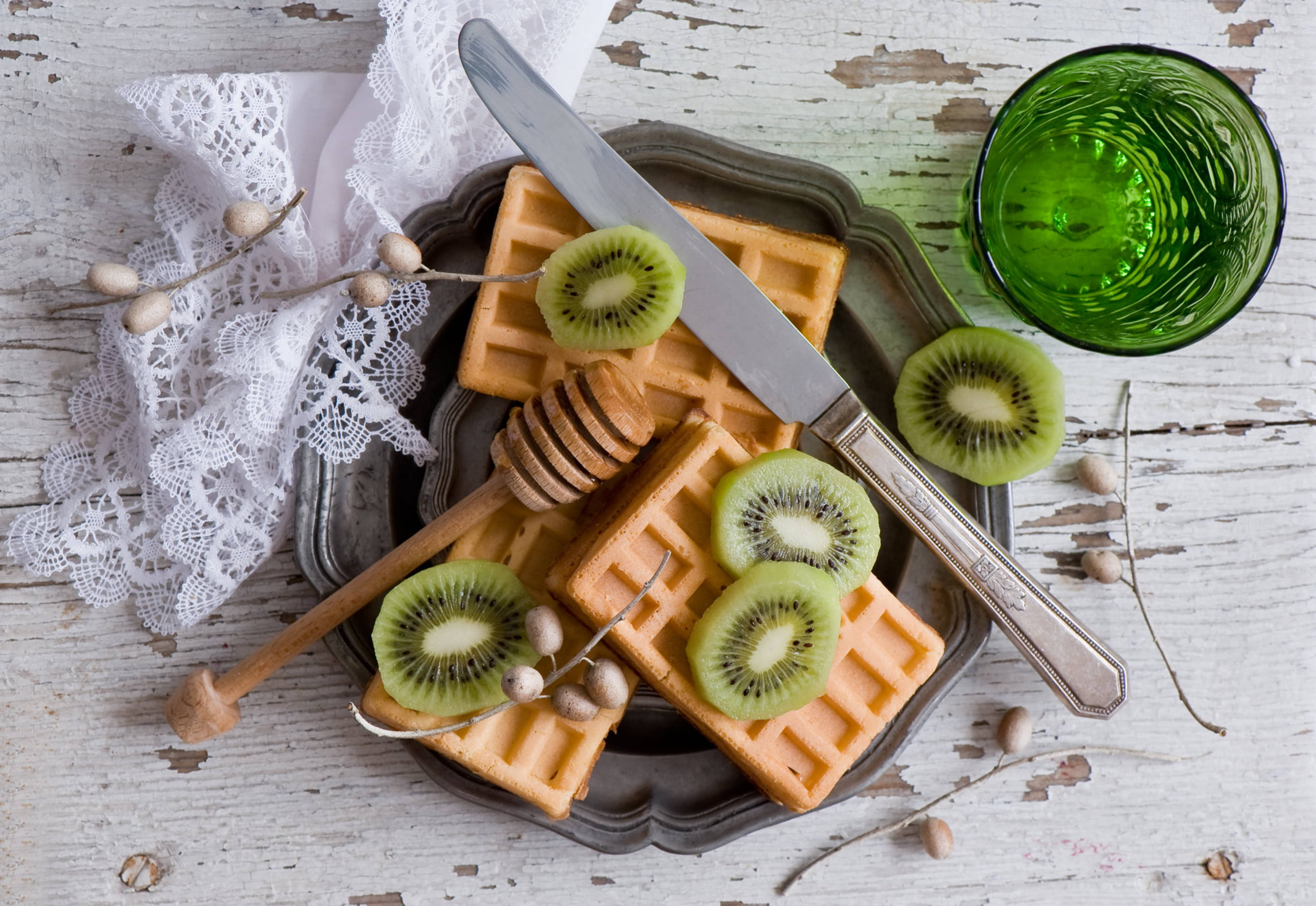 Waffle 桌布 壁紙 — 金黃色鬆餅配上新鮮奇異果片和蜂蜜蘸棒，放在復古托盤上，旁邊有一個綠色玻璃杯，置於鄉村風白色木桌上。
