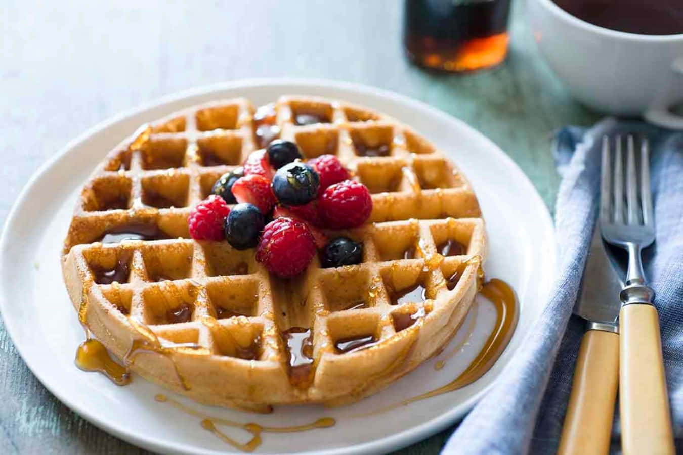 Waffle 桌布 壁紙 — 金黃色鬆餅配上新鮮莓果和糖漿，放在白色盤子上，旁邊有杯子和餐具。