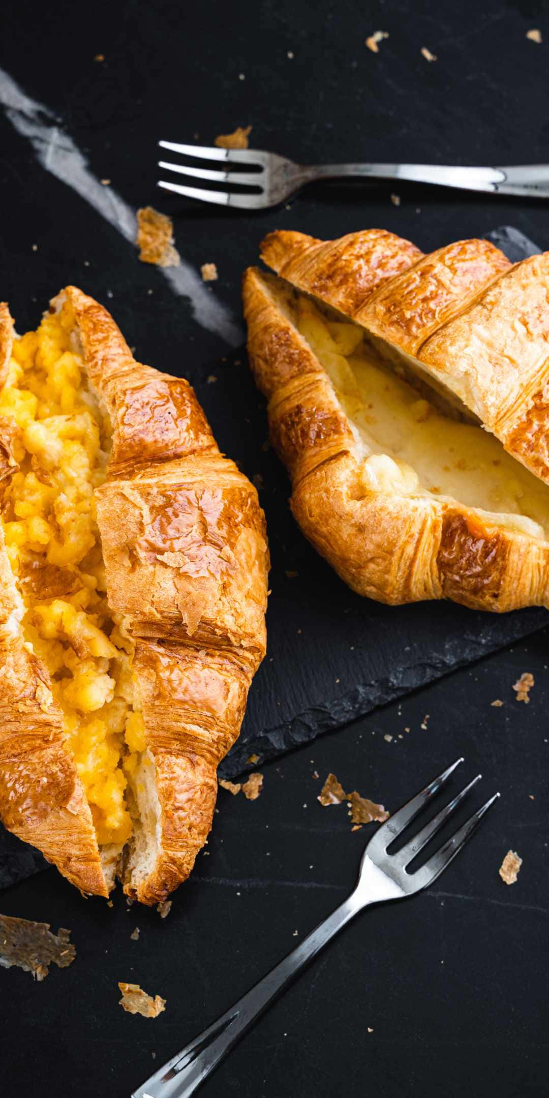 Pastries 桌布 壁紙 — 金黃酥脆的可頌內填滿奶油炒蛋，放在深色石板上的散落麵包屑和叉子之間。