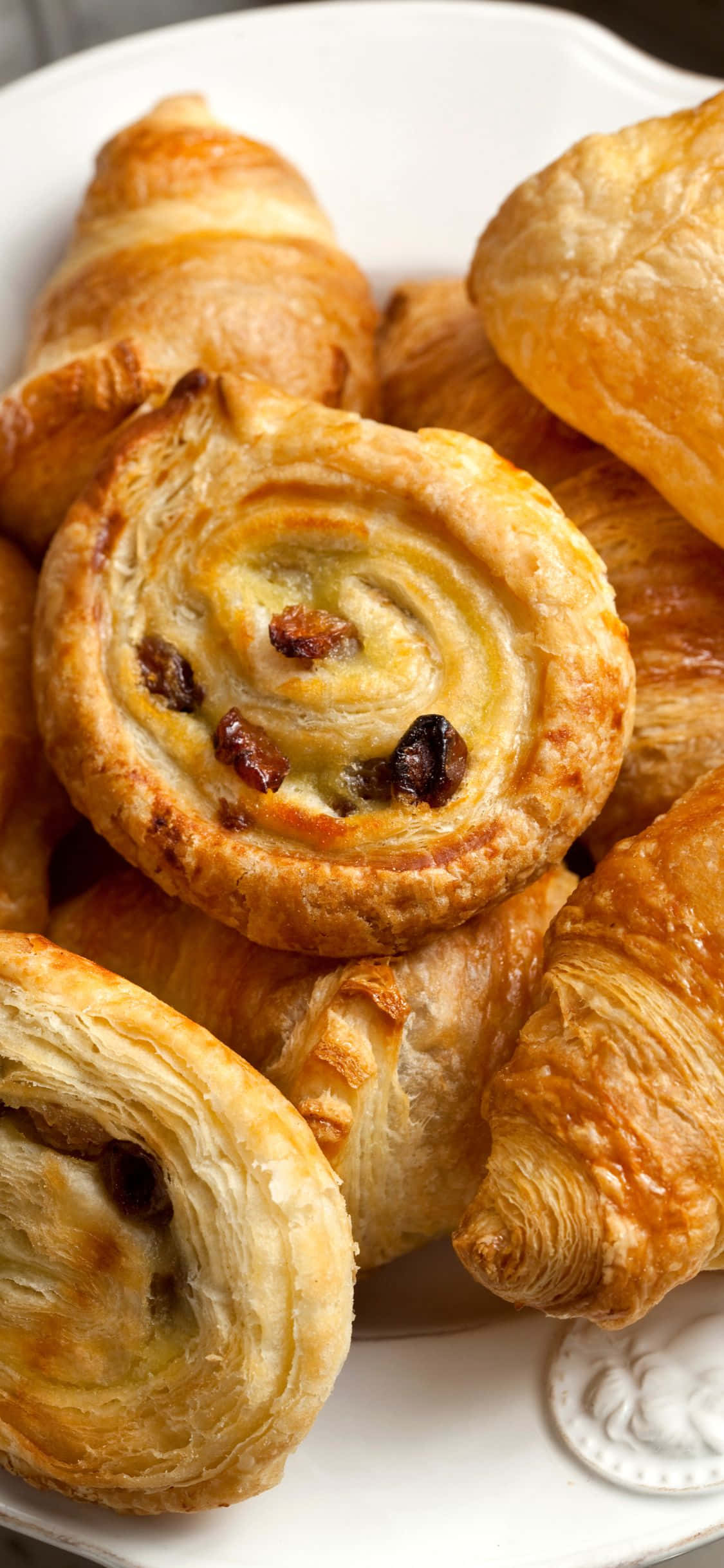 iPhone Xs Pastries 桌布 壁紙 — 金黃酥脆的可頌和葡萄乾漩渦糕點高高堆疊在白色盤子上。