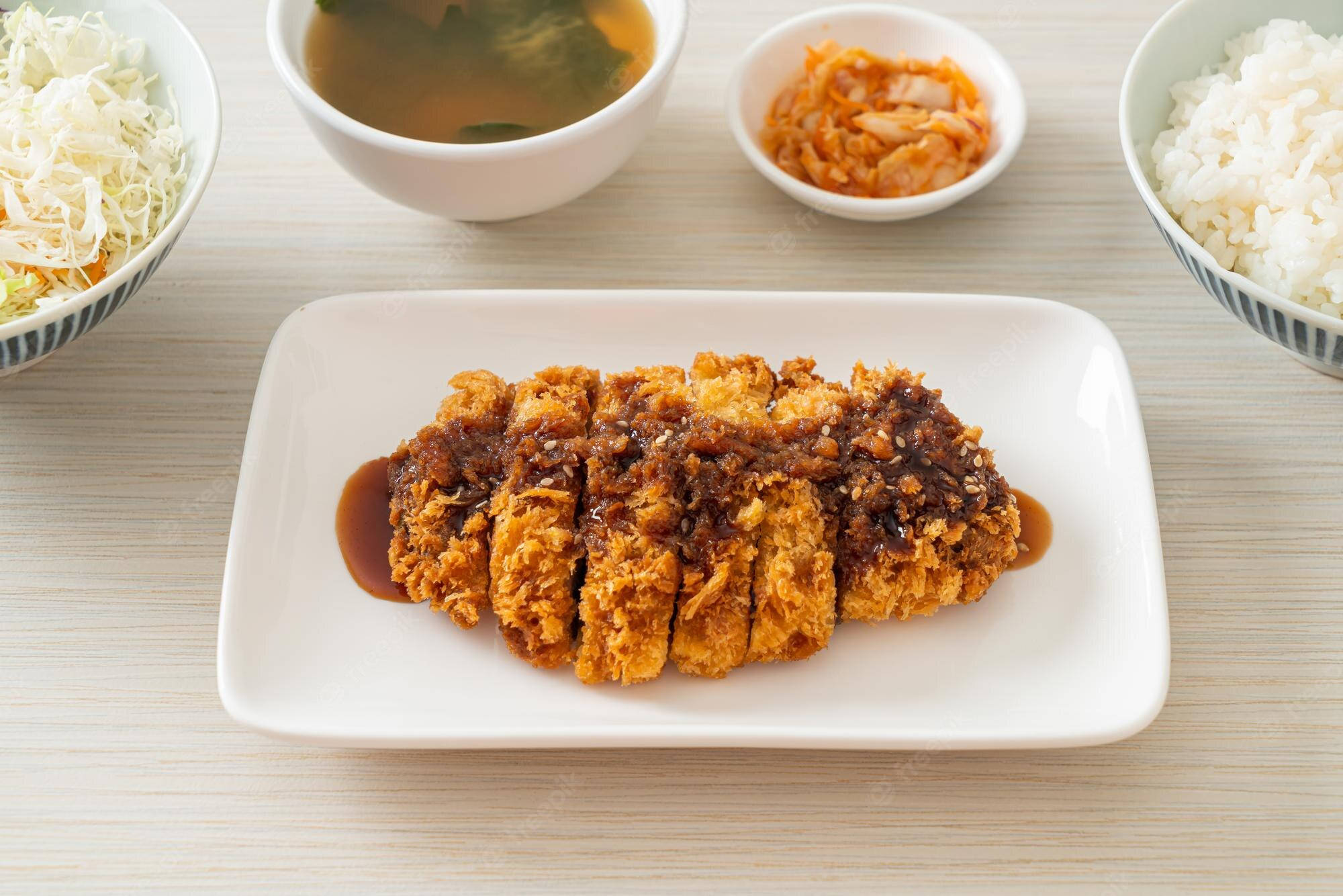 Food Photography 桌布 壁紙 — 金黃酥脆的吉列豬排淋上濃郁醬汁，放在淺色木桌中央的盤中，周圍環繞著米飯、泡菜湯和高麗菜絲。