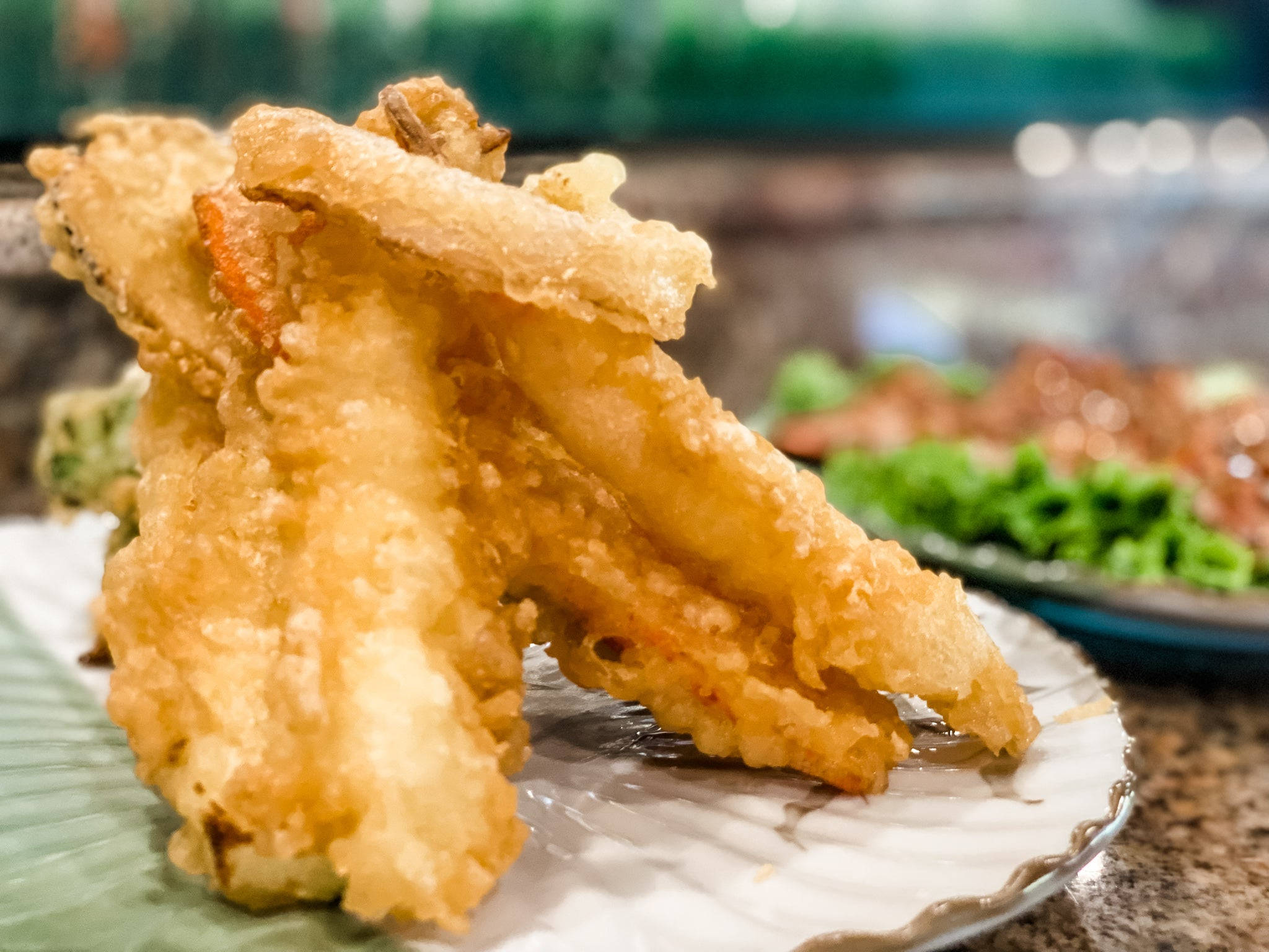 shrimp 桌布 壁紙 — 金黃酥脆的天婦羅蔬菜高高堆在白色盤子上，帶有模糊的綠色裝飾和背景。
