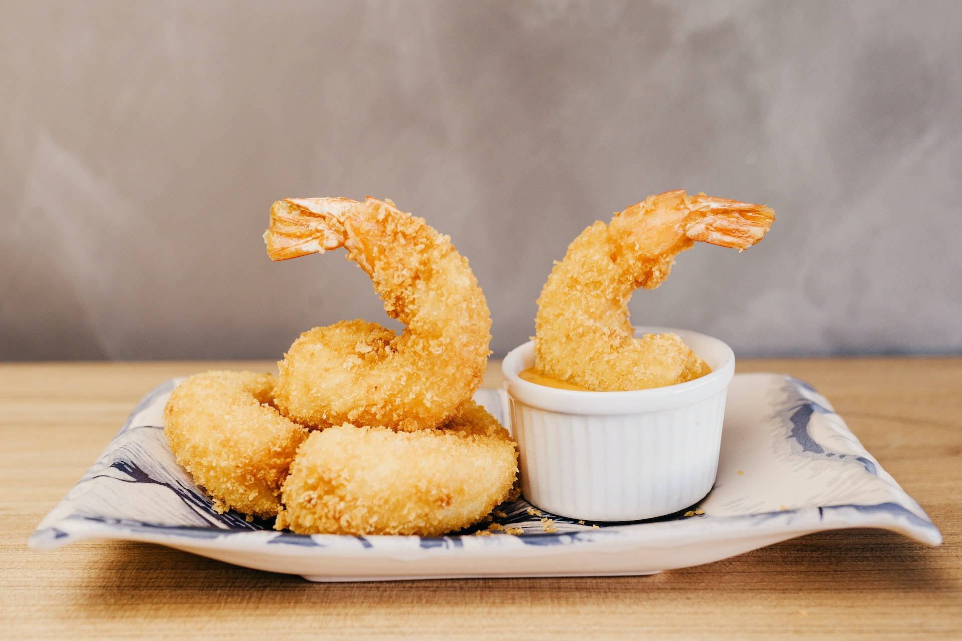 shrimp 桌布 壁紙 — 金黃酥脆的炸蝦堆疊在帶圖案的盤子上，其中一隻蘸了醬汁，背景是柔和的灰色。