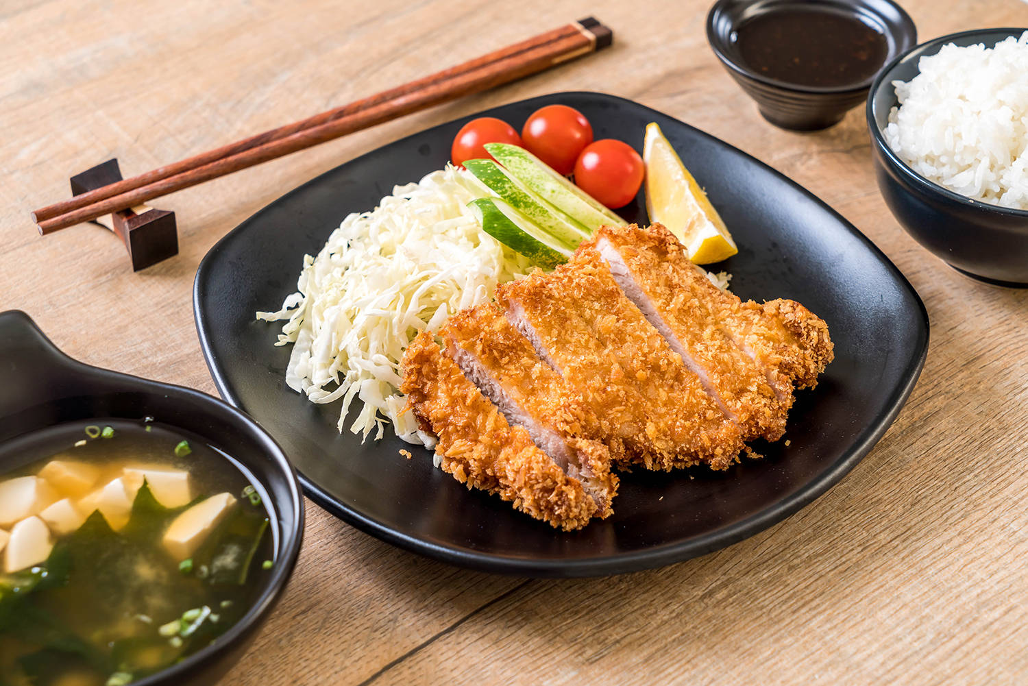 Food Photography 桌布 壁紙 — 金黃酥脆的炸豬排放在黑色盤子上，搭配米飯、醃菜、番茄和湯，置於木桌上。