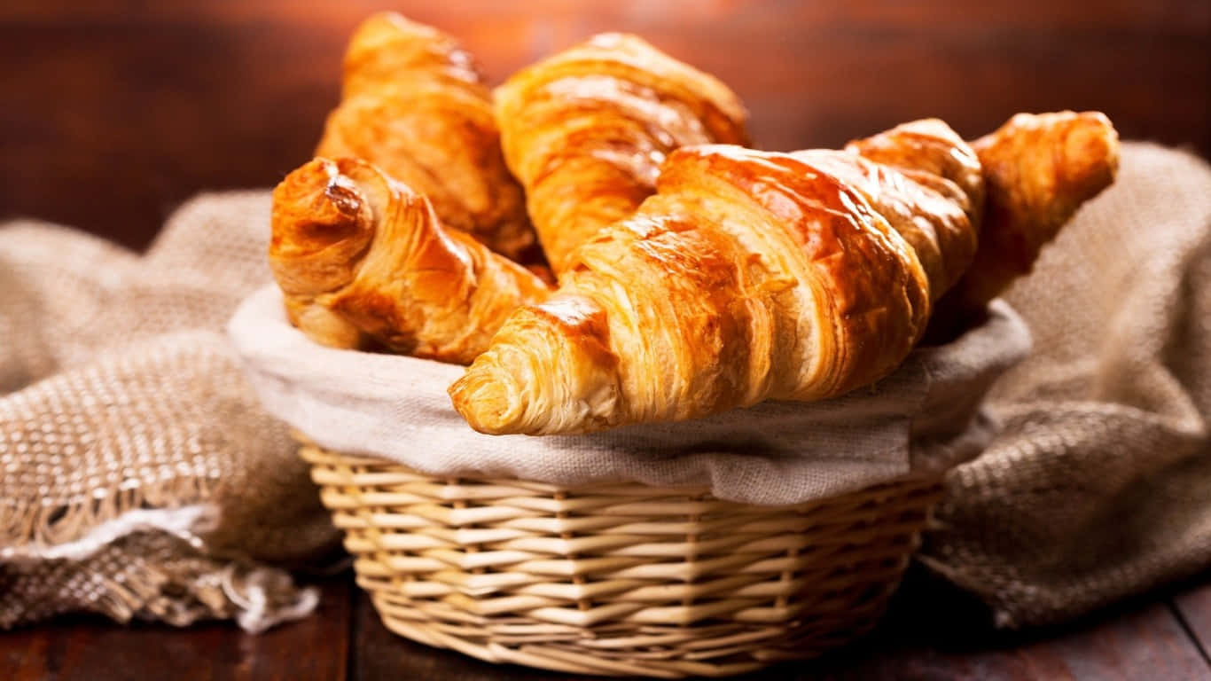 1366x768 Pastries 桌布 壁紙 — 金黃酥脆的牛角麵包放在鋪有布的編織籃子裡，置於質樸的木桌上。