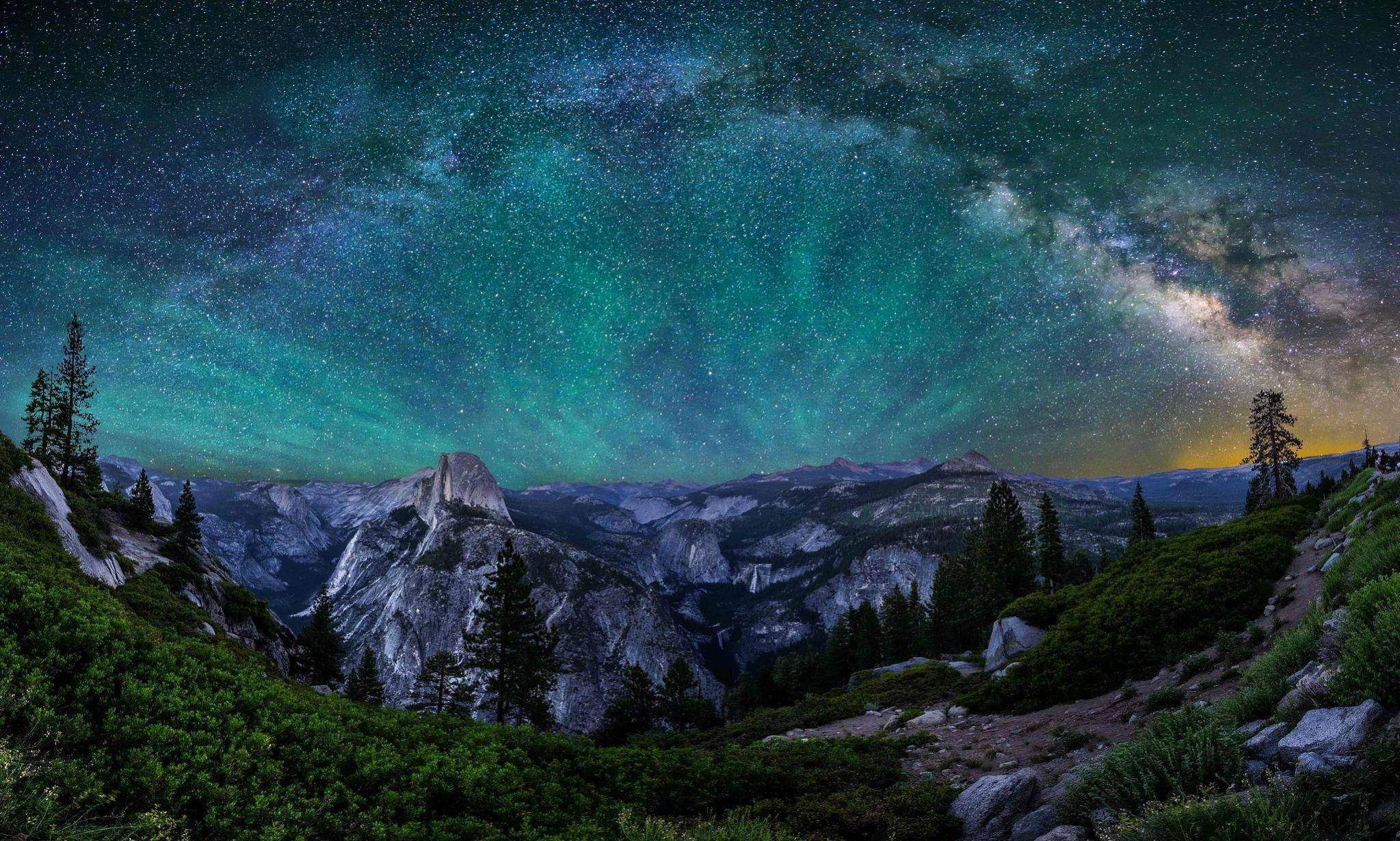 夜空 桌布 壁紙 — 銀河在Half Dome上空拱起，下方是松樹和岩石地形，夜空呈現青綠色和紫色，繁星點點。