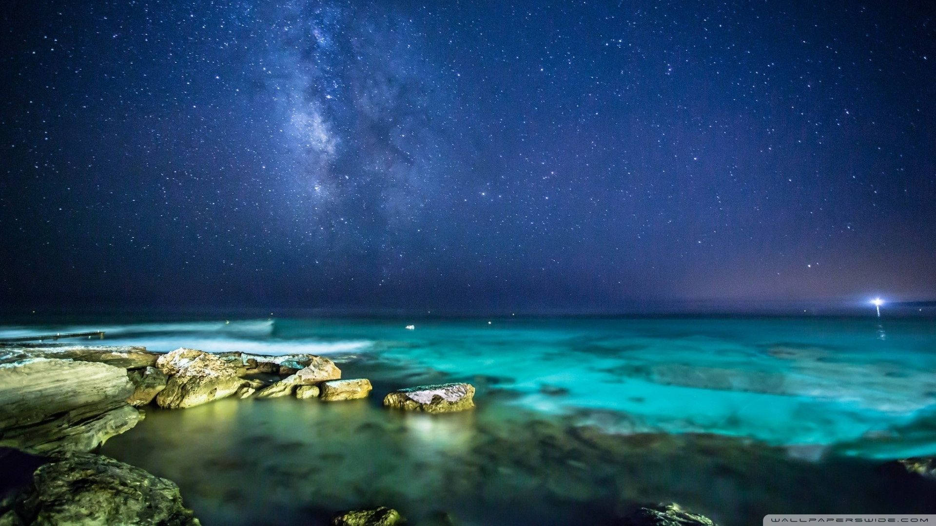 夜空 桌布 壁紙 — 銀河在繁星點點的夜空中拱起，下方是閃爍的藍綠色海浪拍打著黑暗的岩石海岸。
