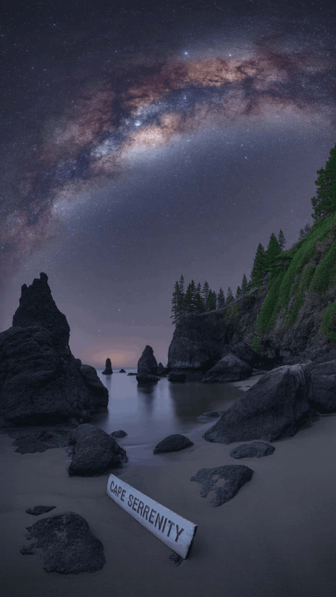 Night sky and seascape at cape serenity milky way wallpaper 壁紙 — 銀河拱起在月光照耀的岩石海灘上，一個寫著Cape Serenity的標誌 nestled among dark boulders and pine trees.