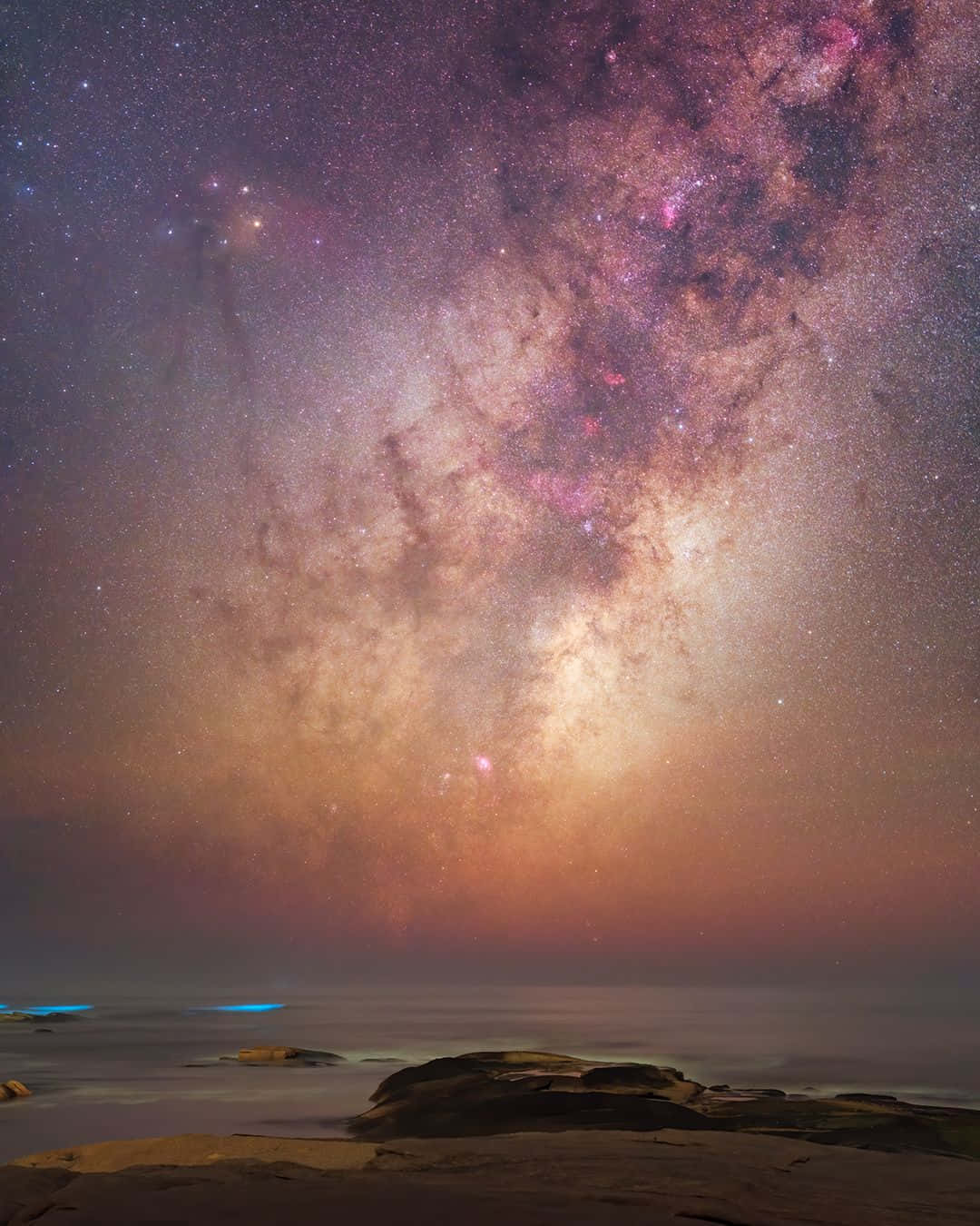 Galactic Center 桌布 壁紙 — 銀河系以鮮豔的紫色和金色弧形橫跨在霧氣瀰漫的海洋上，前景是黑暗的岩石。