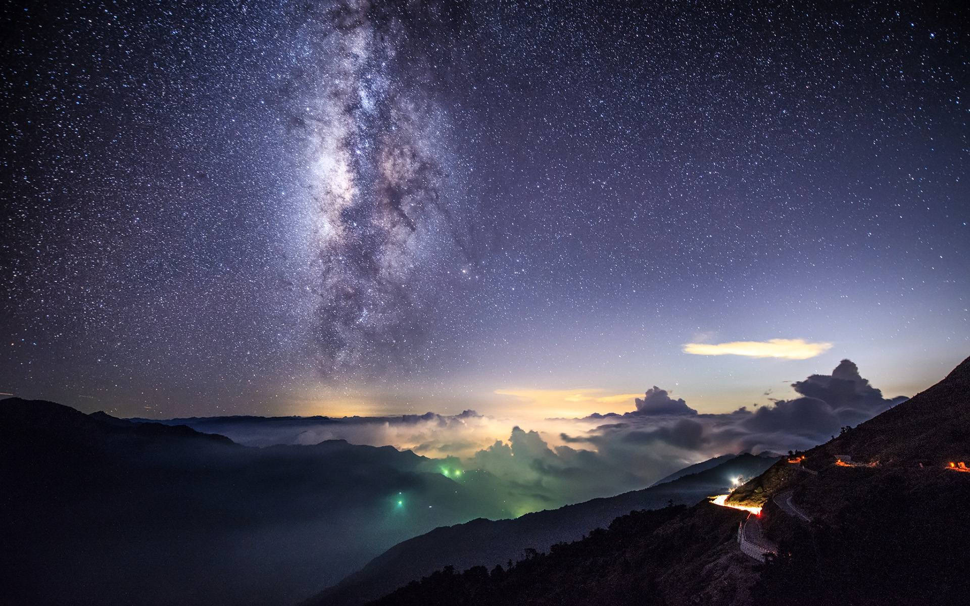 Milky 桌布 壁紙 — 銀河系在山峰上空拱起，雲海之上，有發光的綠色極光和汽車燈光照亮的蜿蜒道路。