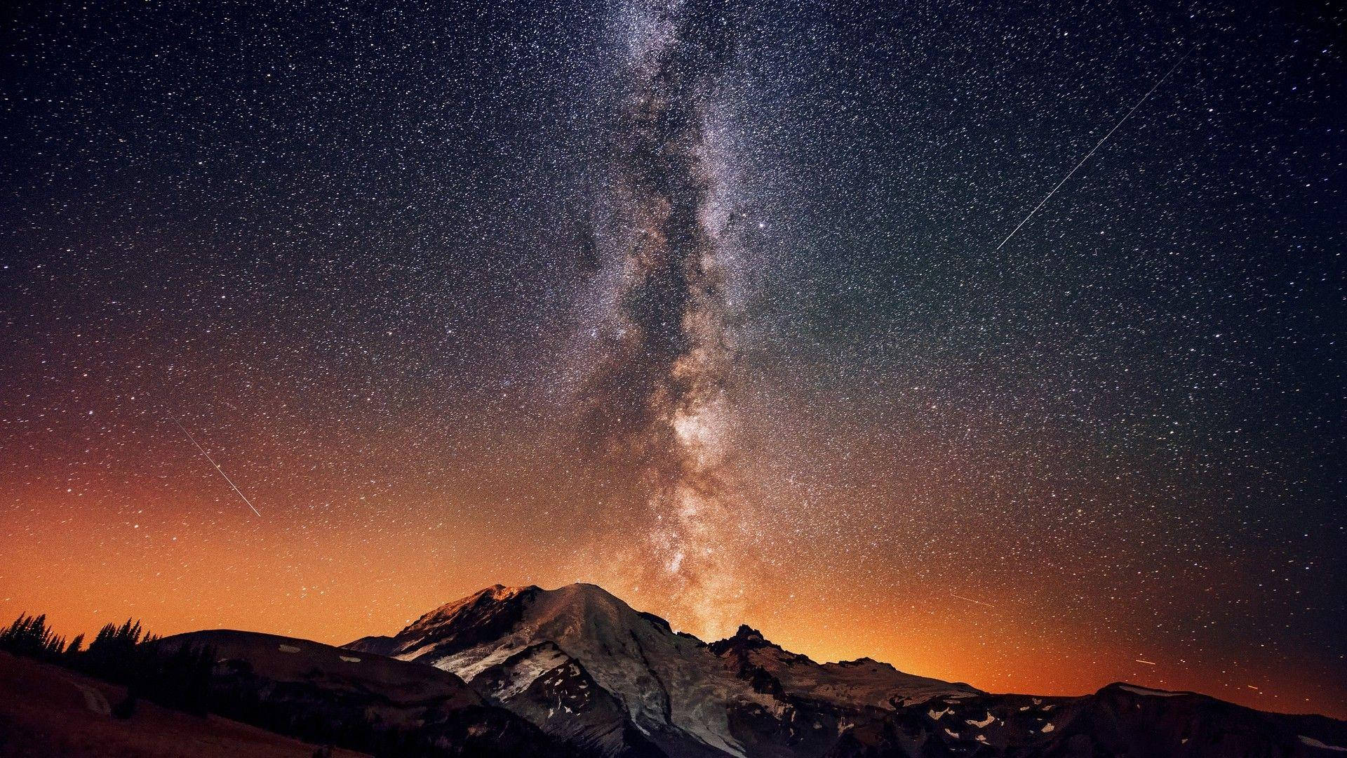 Milky 桌布 壁紙 — 銀河系在星空下戲劇性地拱起，越過積雪的山峰，帶有微弱的橙色光芒。