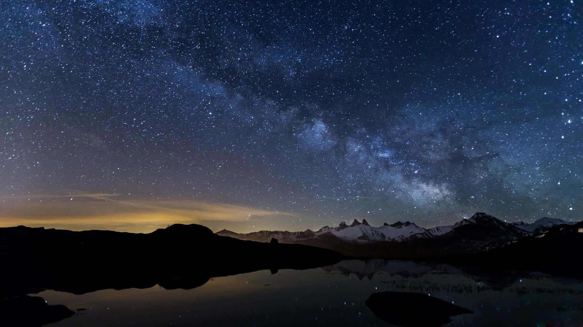 夜空 桌布 壁紙 — 銀河系在積雪的山峰上空劃出一道弧線，倒映在寧靜的高山湖泊中，星空璀璨。
