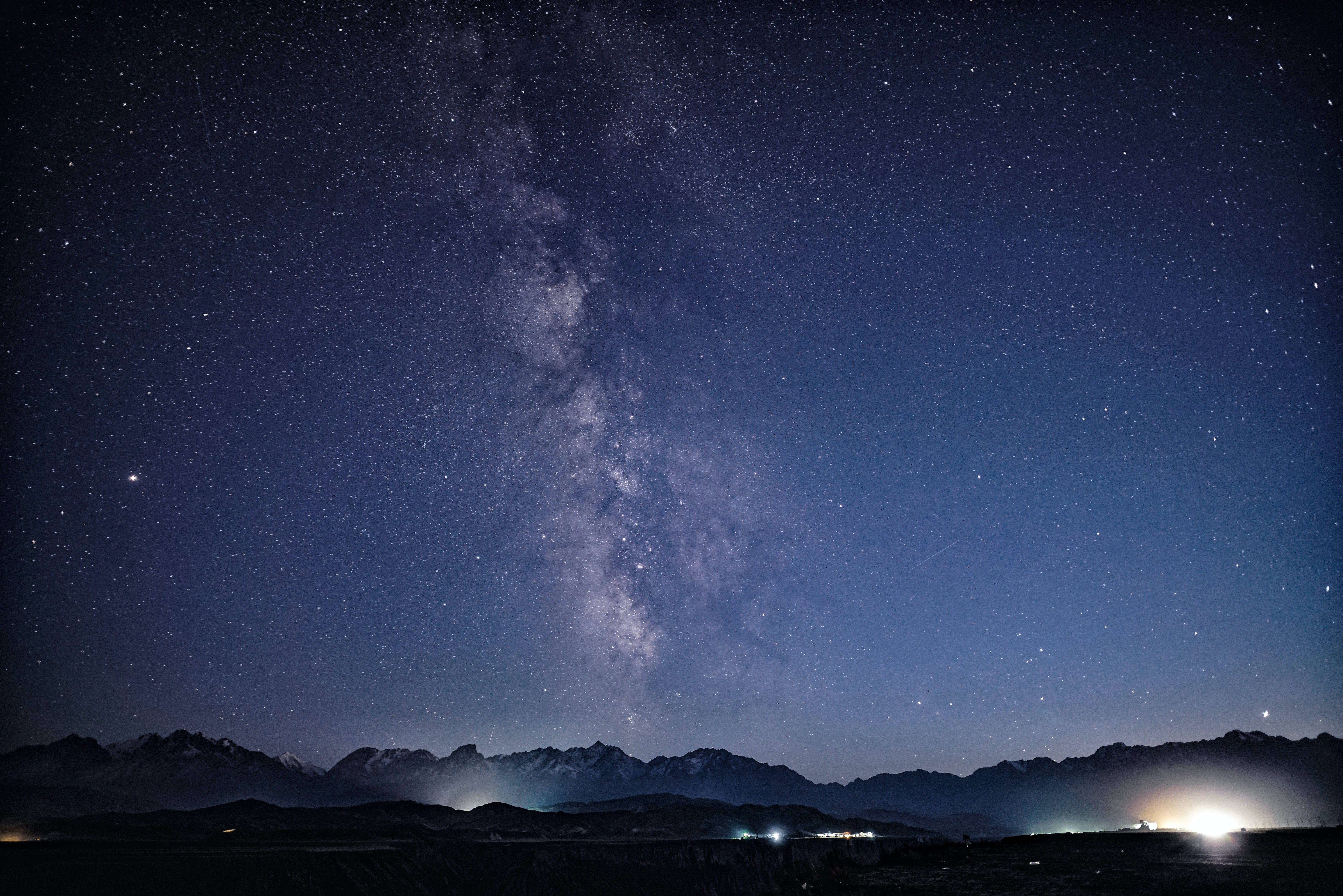夜空 桌布 壁紙 — 銀河系在繁星點點的夜空上，於積雪的山峰之上燦爛地彎曲，下方有遠處的燈光。