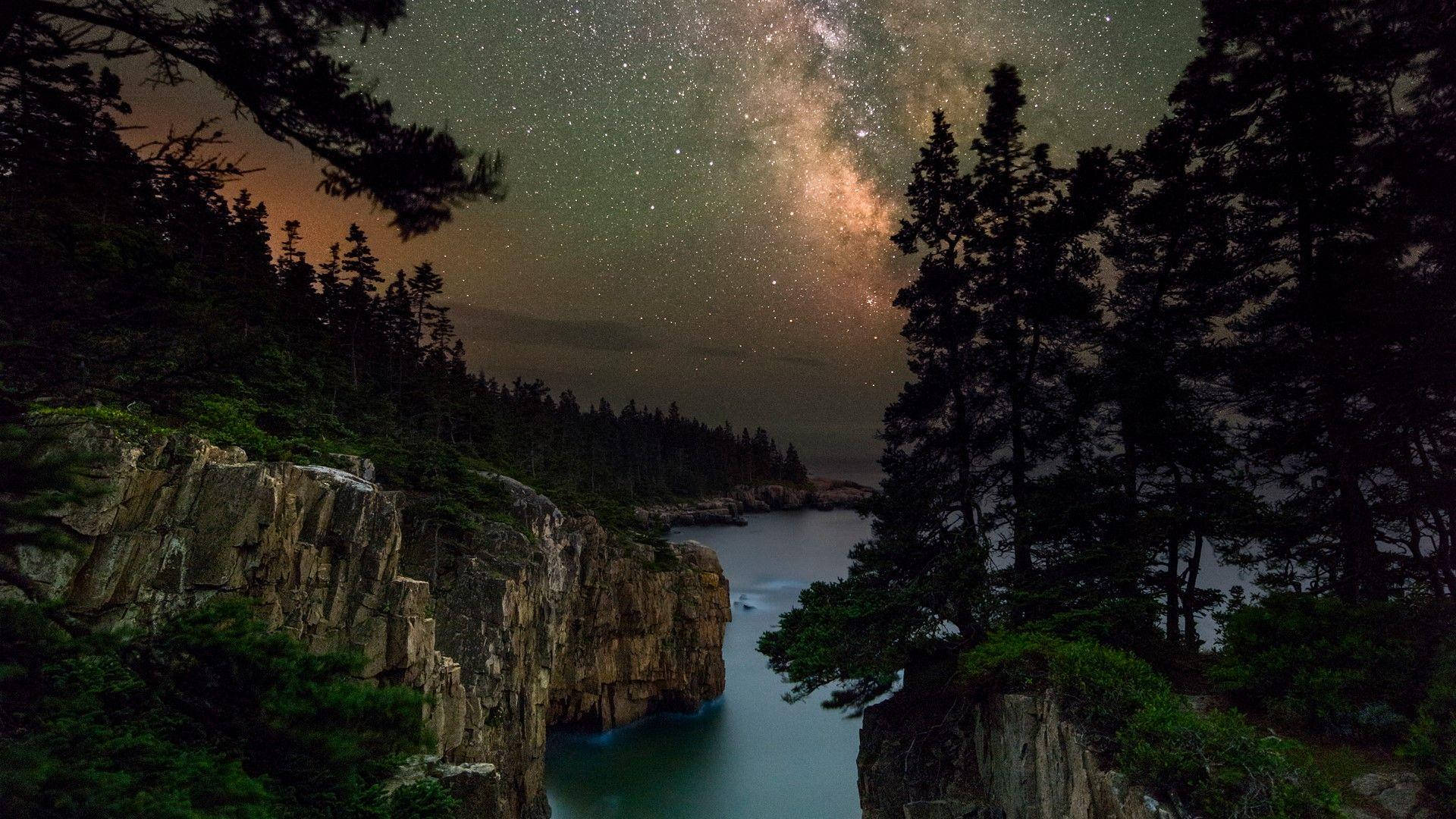 樹木 桌布 壁紙 — 銀河系在繁星點點的夜空上拱起，下方是一個僻靜的小海灣，四周環繞著深色松樹和崎嶇的懸崖。