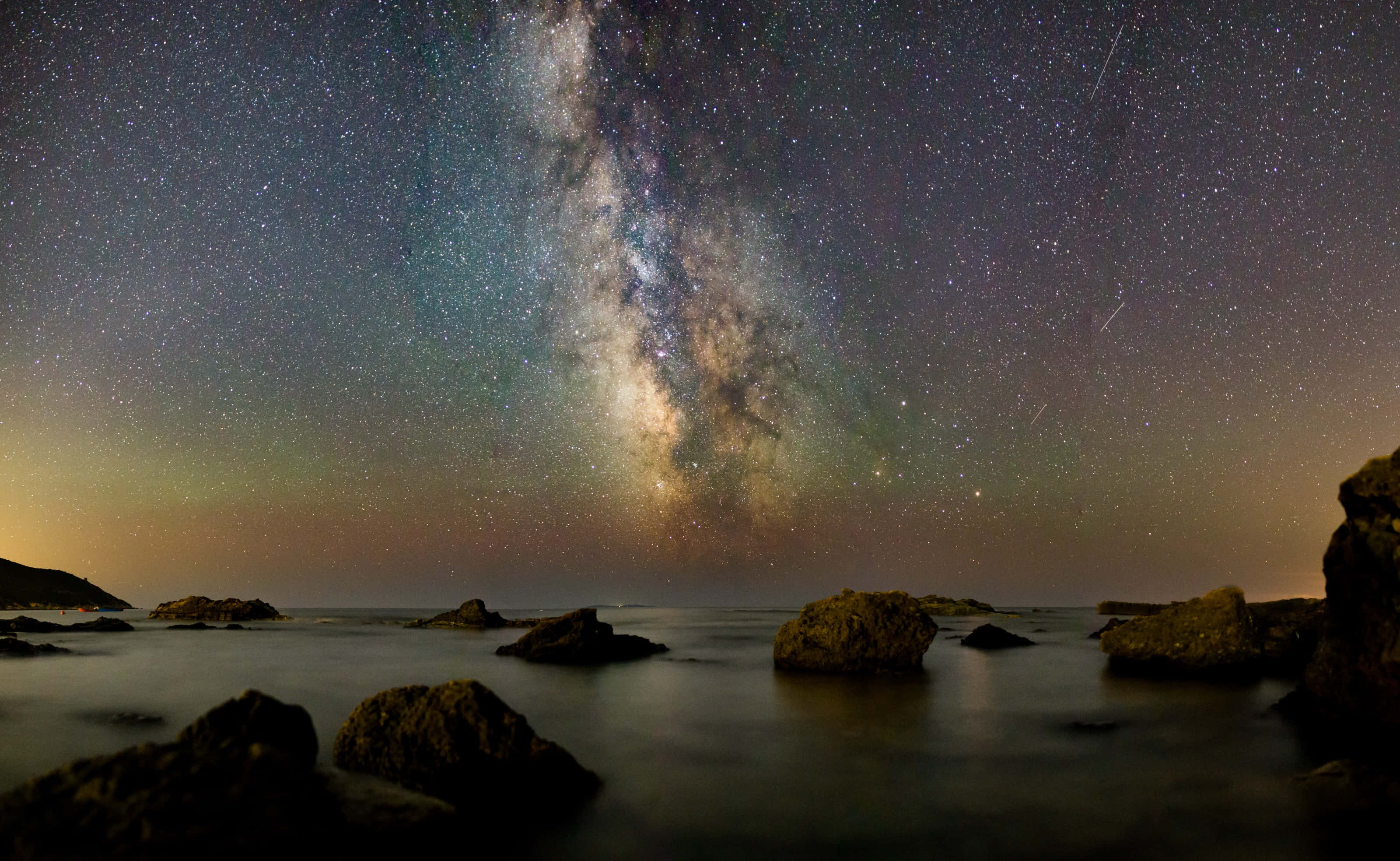 Star Sky 桌布 壁紙 — 銀河系在繁星點點的夜空下，在平靜的海洋和前景的黑色岩石上燦爛地拱起。