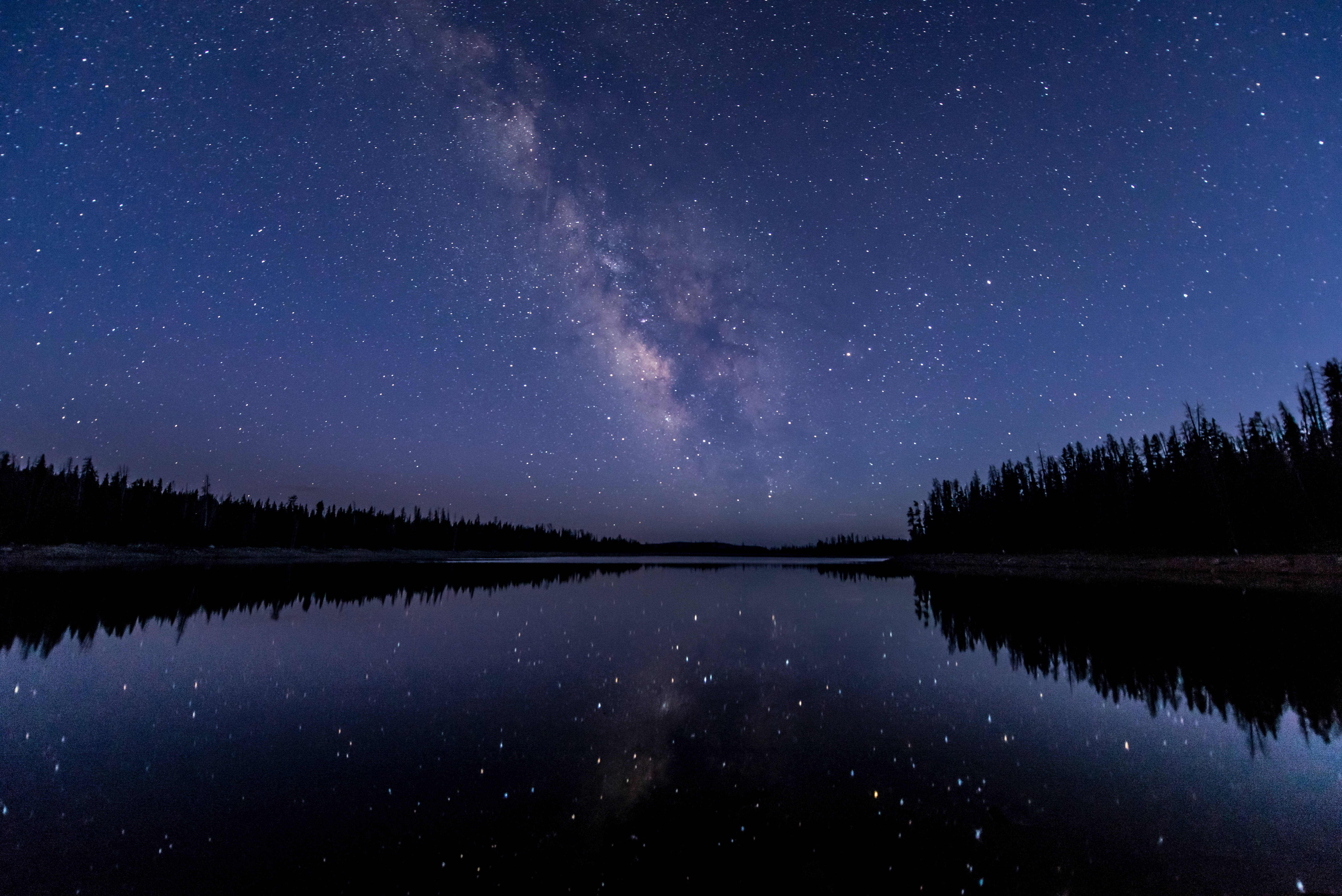 夜空 桌布 壁紙 — 銀河系在繁星點點的夜空中劃出一道弧線，完美倒映在平靜的湖面上，四周環繞著黑暗的森林剪影。