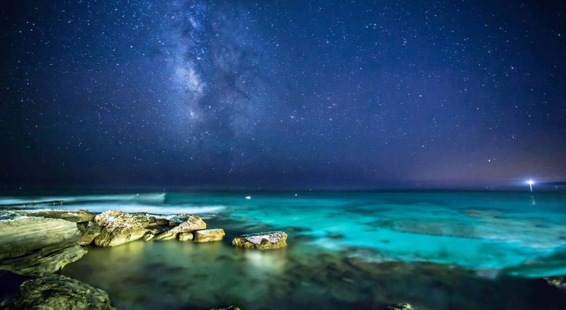 Stars 桌布 壁紙 — 銀河系在繁星點點的夜空中拱起，下方是碧綠色的海浪拍打岩石海岸。