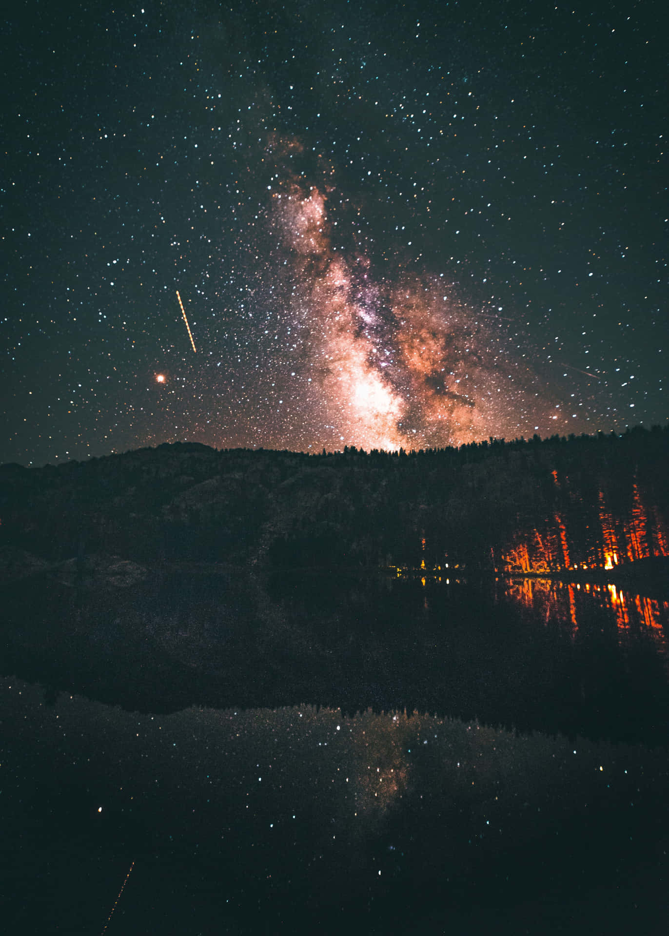 Night Aesthetic 桌布 壁紙 — 銀河系在繁星點點的夜空中拱起，倒映在平靜的湖面上，岸邊有發光的橙色樹木。