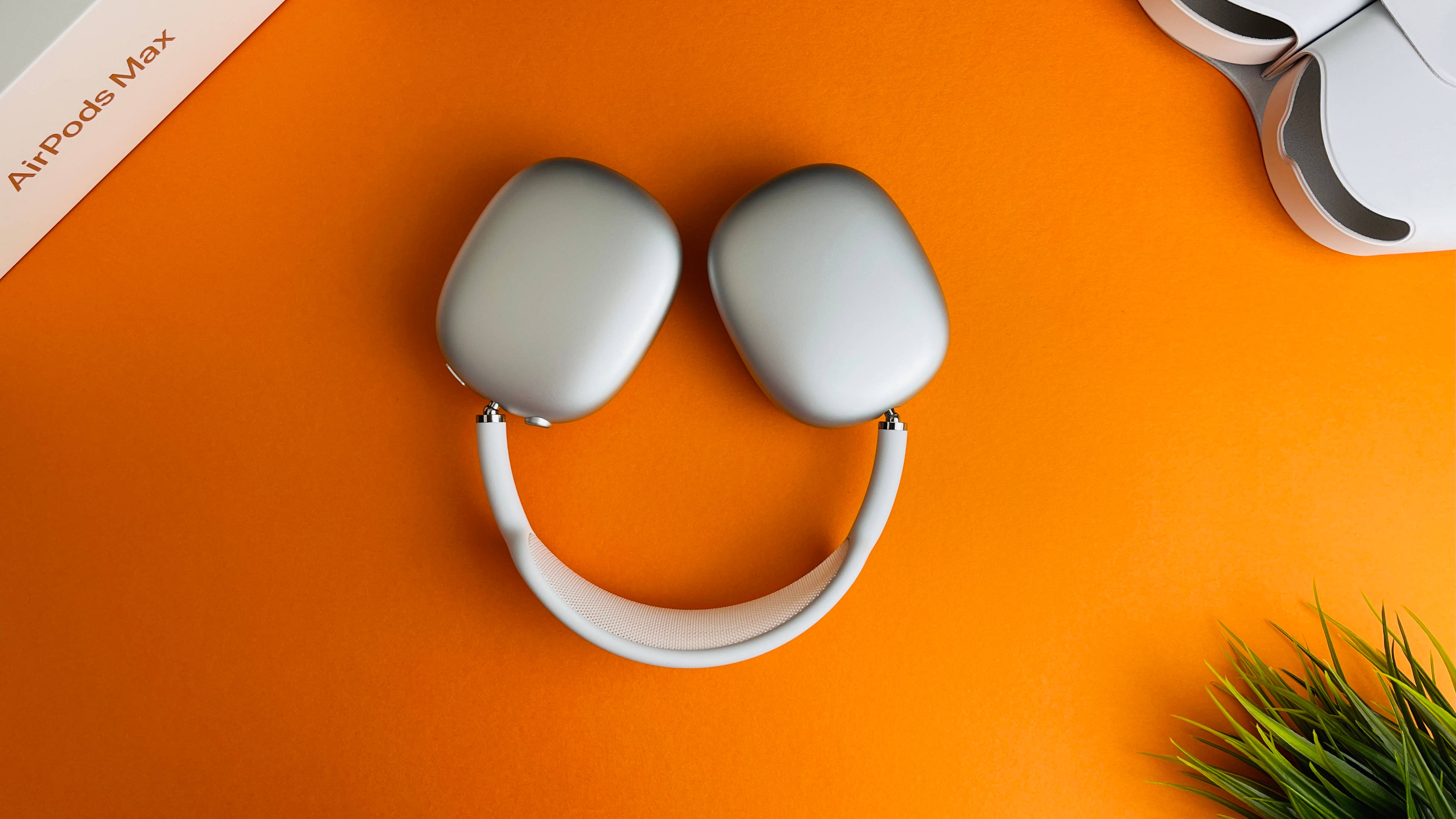 橘色美學 桌布 壁紙 — 銀色 AirPods Max 耳機以笑臉形式排列在充滿活力的橙色背景上，配有綠色植物和包裝。