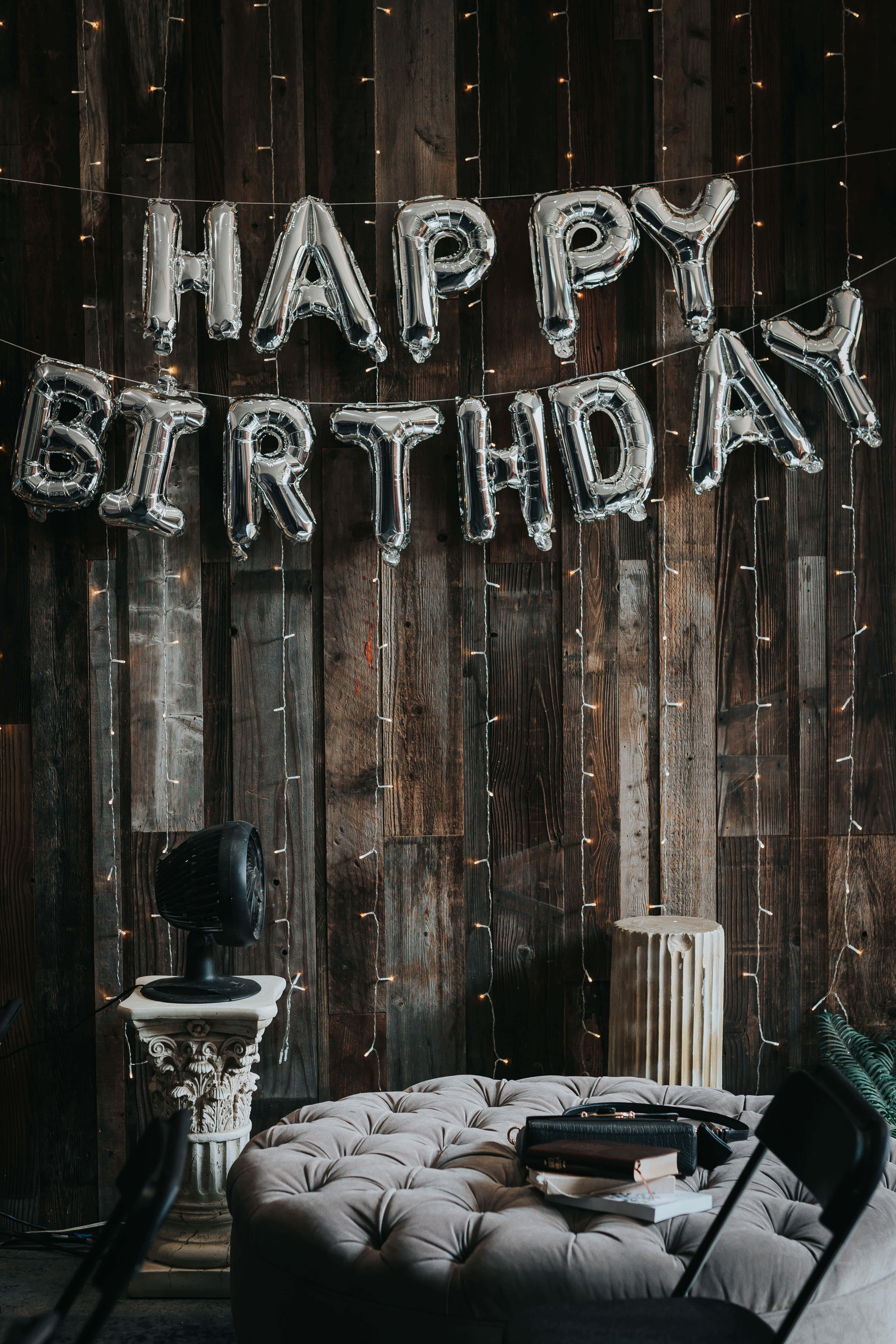 High Resolution Happy Birthday 桌布 壁紙 — 銀色 'HAPPY BIRTHDAY' 氣球掛在質樸的木牆上，牆上裝飾著燈串，下方是簇絨灰色軟凳。