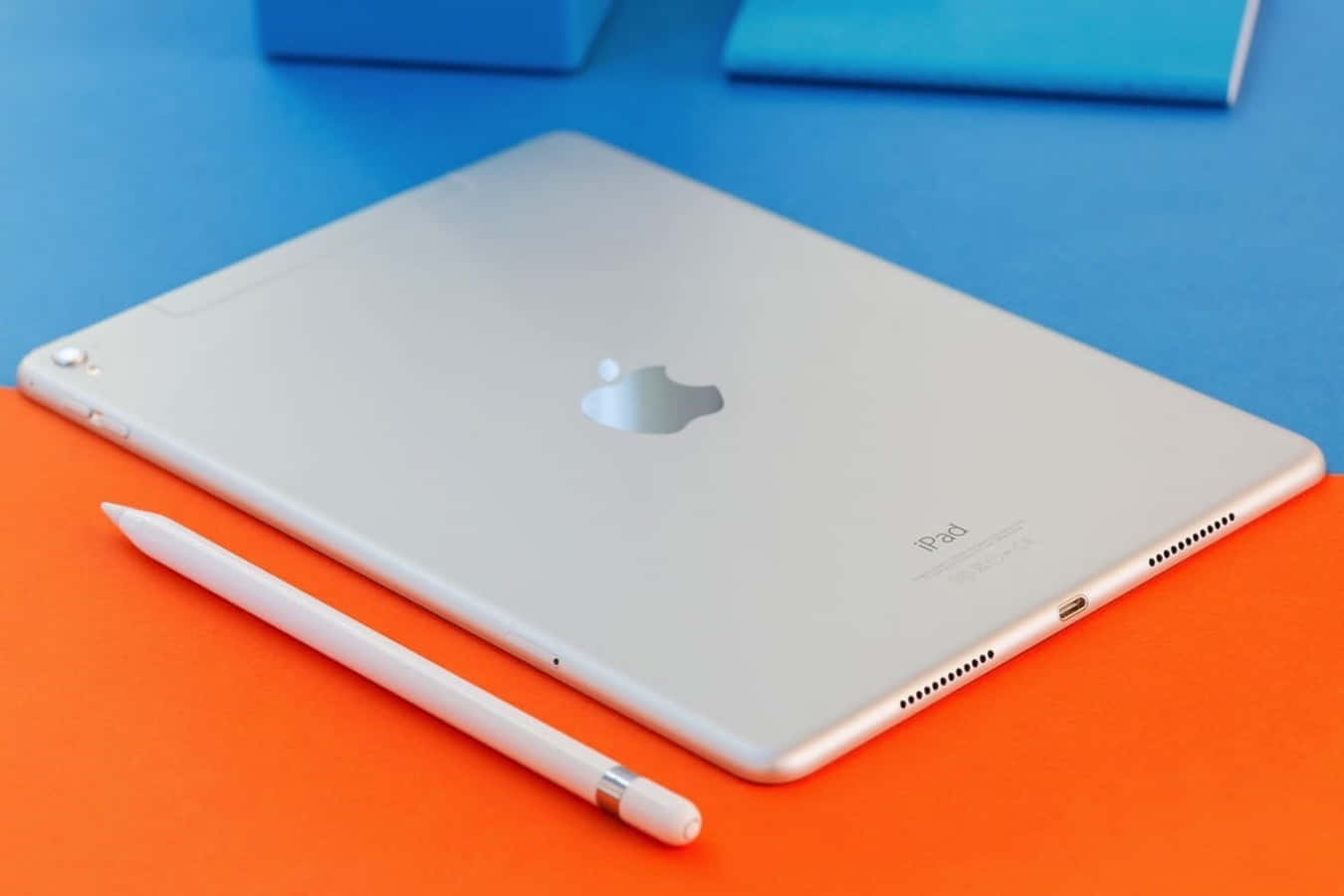 iPad 桌布 壁紙 — 銀色 iPad 帶有 Apple 標誌，放在鮮橙色表面上，旁邊有白色觸控筆，背景為藍色。