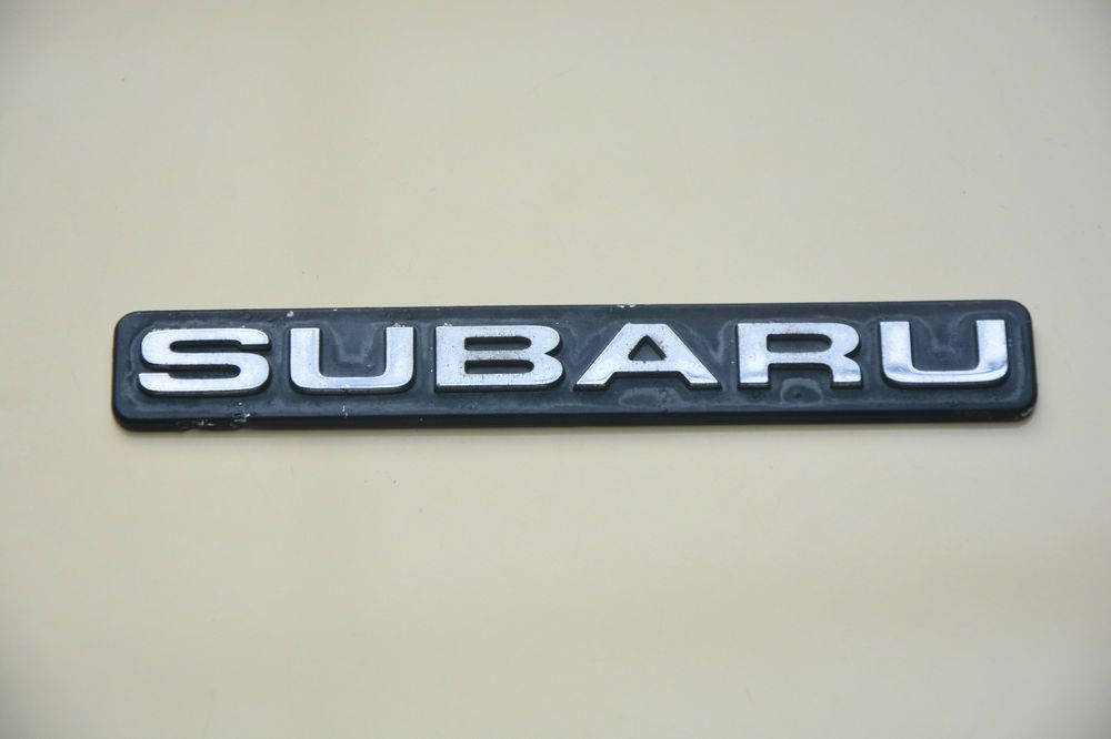 Subaru Logo 桌布 壁紙 — 銀色 Subaru 標誌在米色引擎蓋上的特寫，光線柔和，表面有輕微磨損。