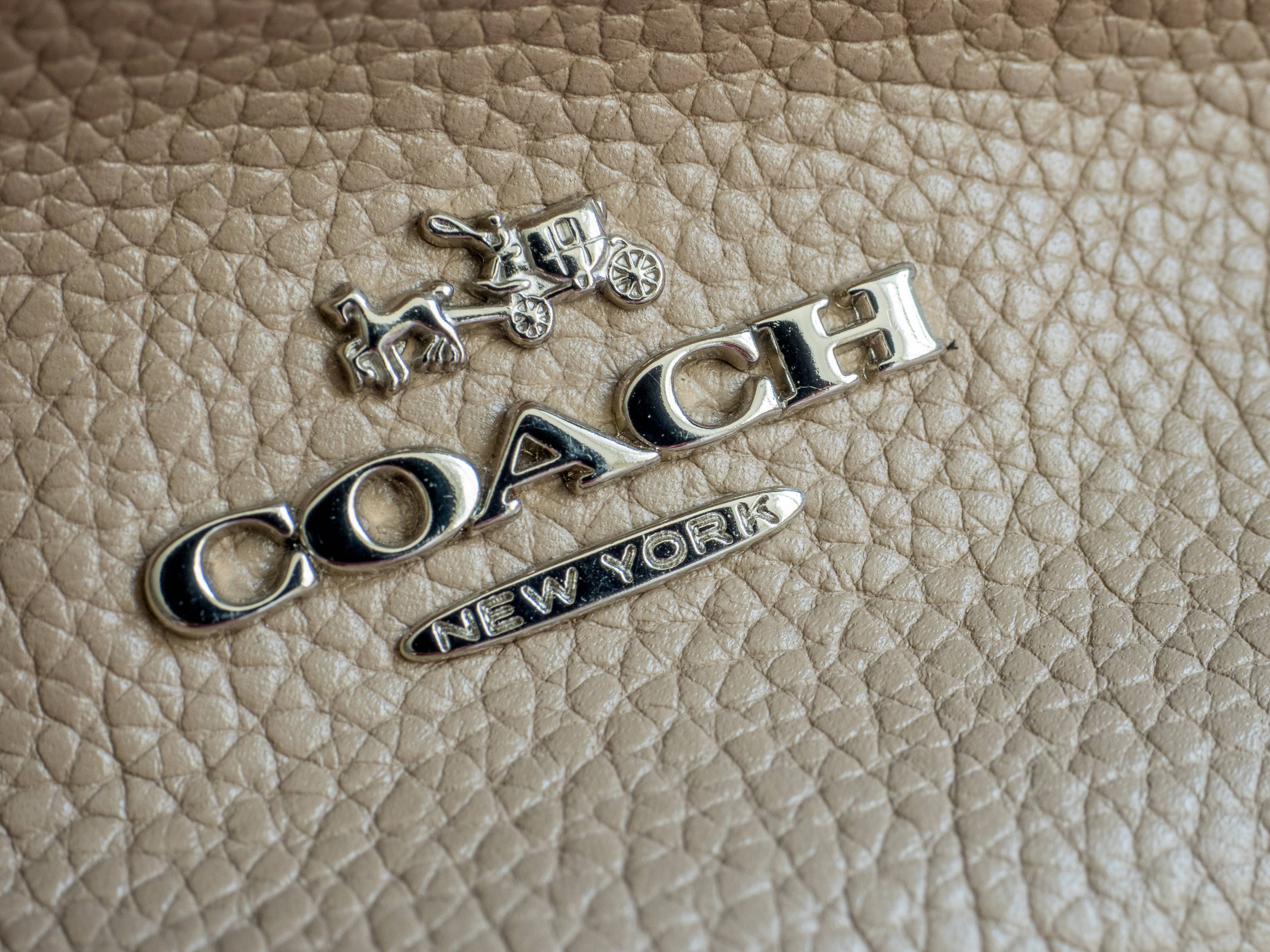 Coach 桌布 壁紙 — 銀色Coach標誌近距離特寫，帶有馬車圖案，位於紋理米色皮革上。