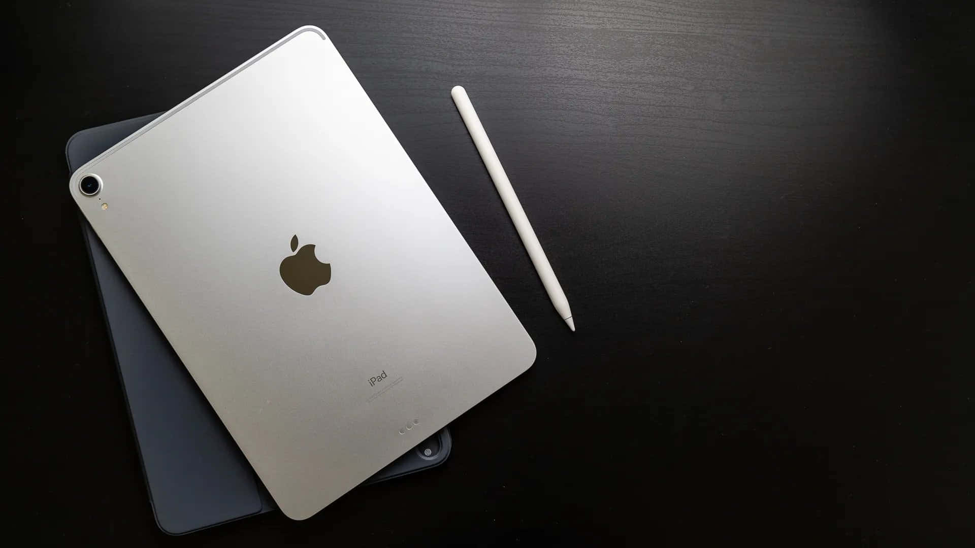 iPad 桌布 壁紙 — 銀色iPad帶有Apple標誌，放在黑色木質表面上的深灰色保護殼上，旁邊有一支白色Apple Pencil。