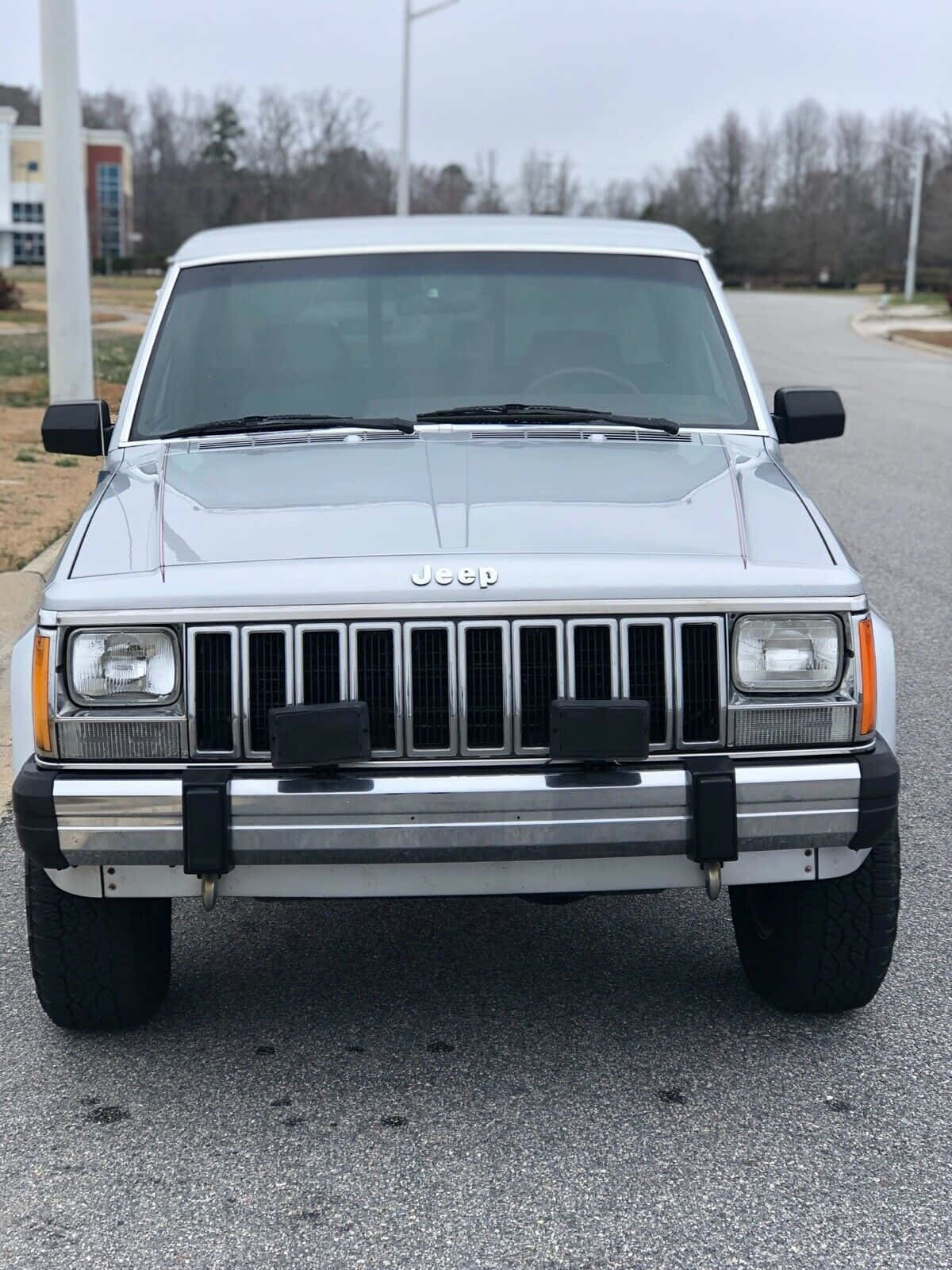Jeep Comanche 桌布 壁紙 — 銀色Jeep Cherokee正面視圖，停在瀝青路上，背景有光禿禿的樹木和陰天。