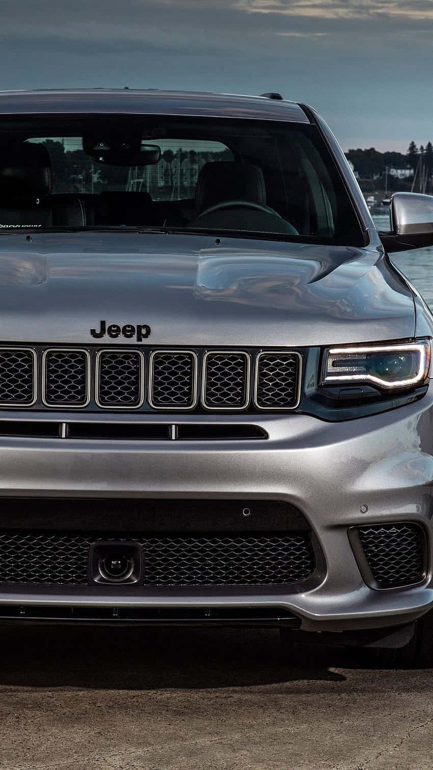 Jeep Trackhawk 桌布 壁紙 — 銀色Jeep SUV的前格柵和頭燈特寫，背景為多雲的天空和海濱。