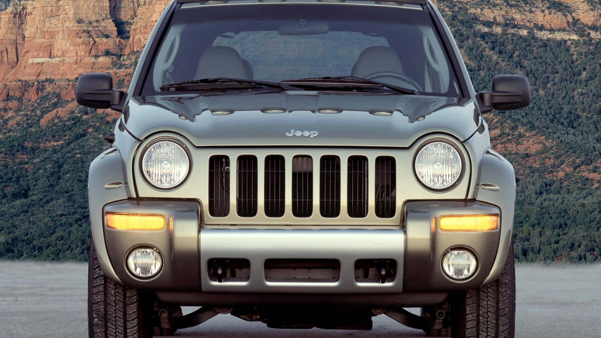 Jeep Liberty 桌布 壁紙 — 銀色Jeep Wrangler停在紅色岩石懸崖和綠色森林前面的正面視圖。