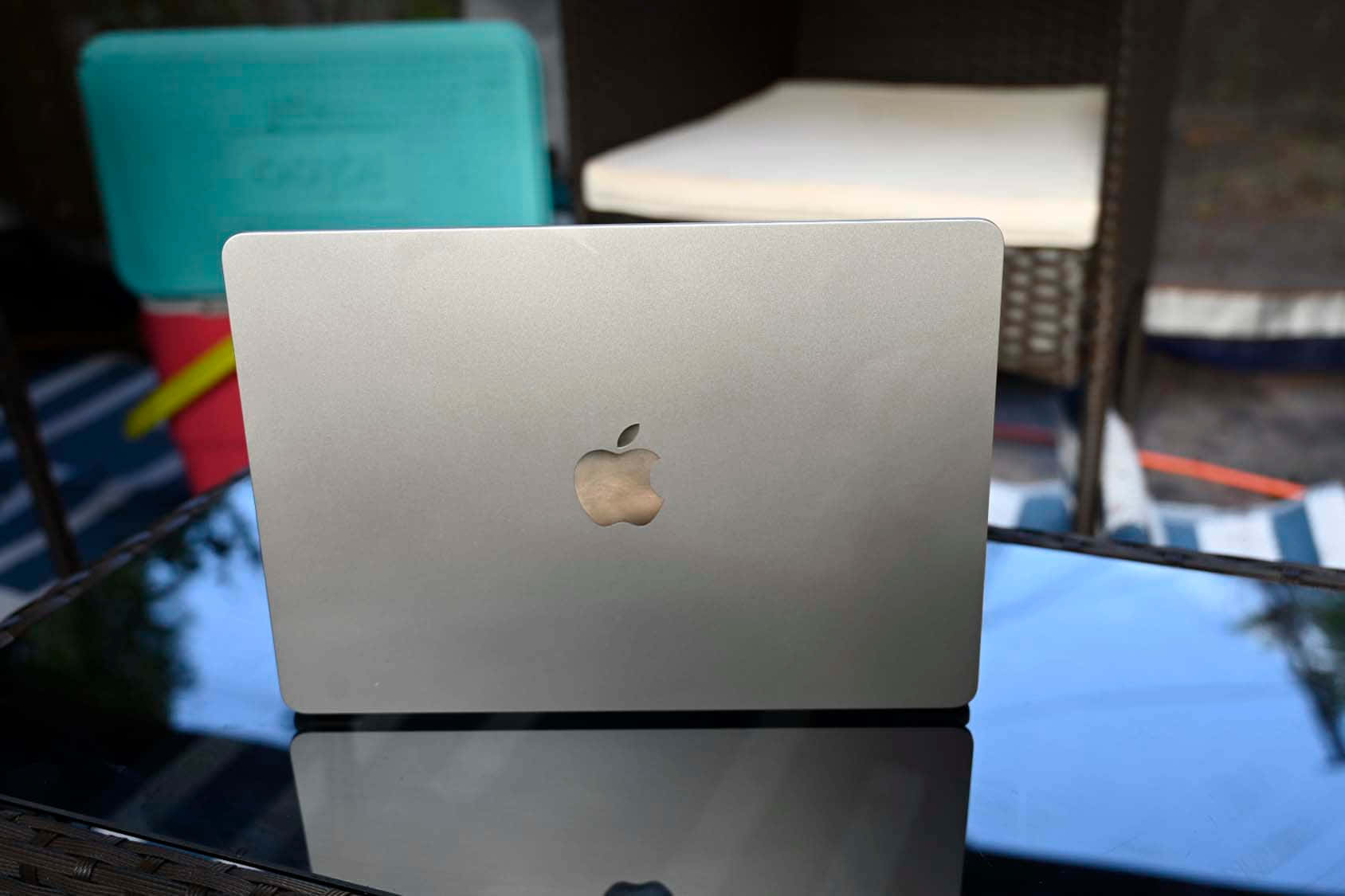 筆電 桌布 壁紙 — 銀色MacBook蓋上帶有蘋果標誌，放在玻璃桌上，反射藍天和戶外庭院家具。