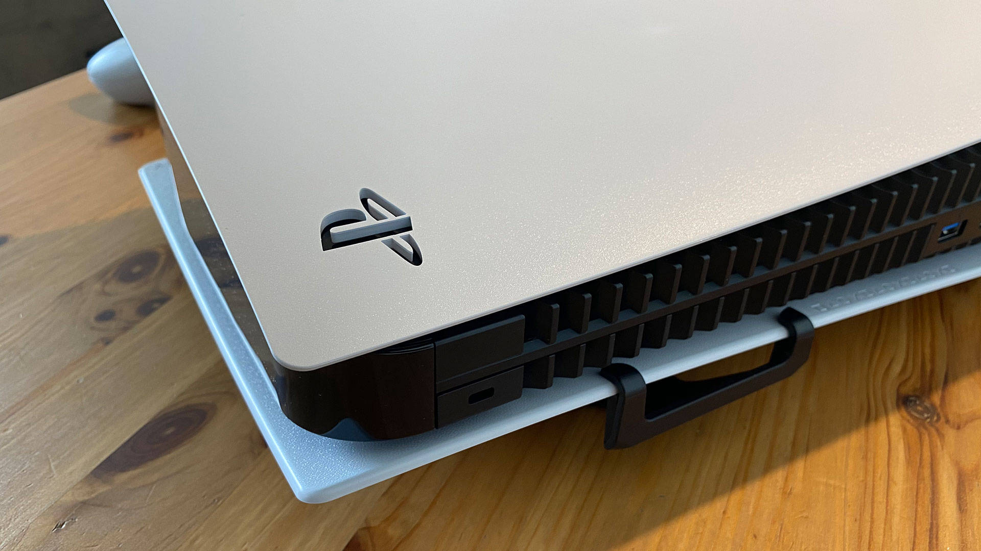 PS5 動態圖片 桌布 壁紙 — 銀色PlayStation 5主機特寫，放置於淺藍色底座上，底座置於溫暖的木桌上。