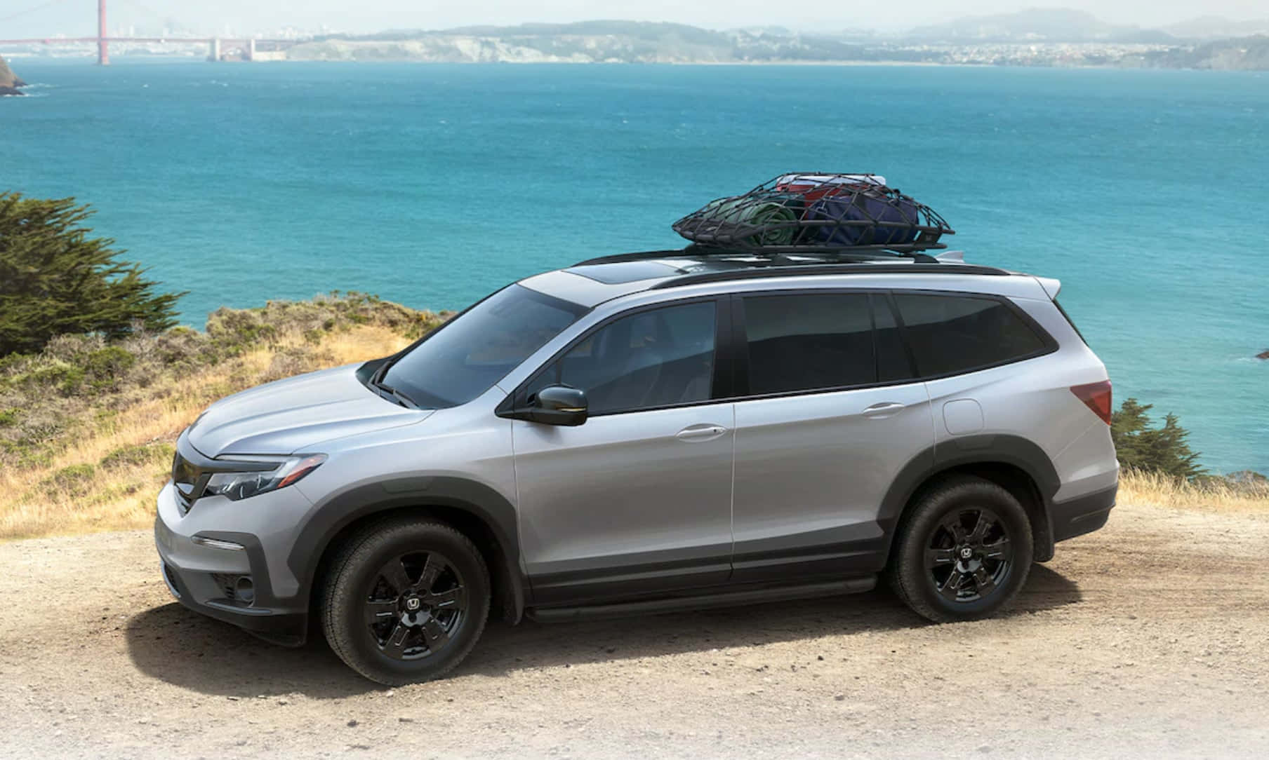 Honda Pilot 桌布 壁紙 — 銀色SUV配備車頂行李架，停泊在懸崖邊，俯瞰碧綠海灣，遠處可見金門大橋。