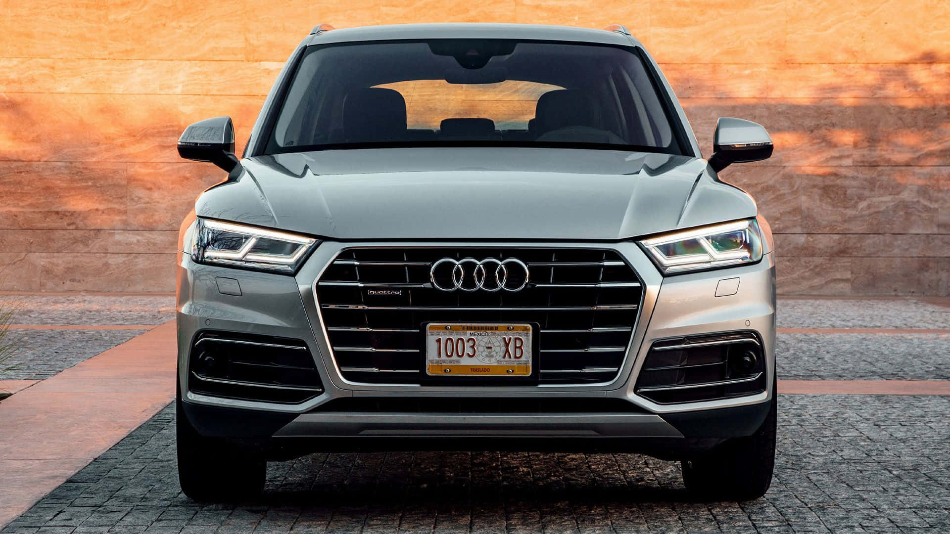 Audi Q5 桌布 壁紙 — 銀色奧迪Q5 SUV正面視圖，停放在鵝卵石車道上，背景為暖色調磚牆。