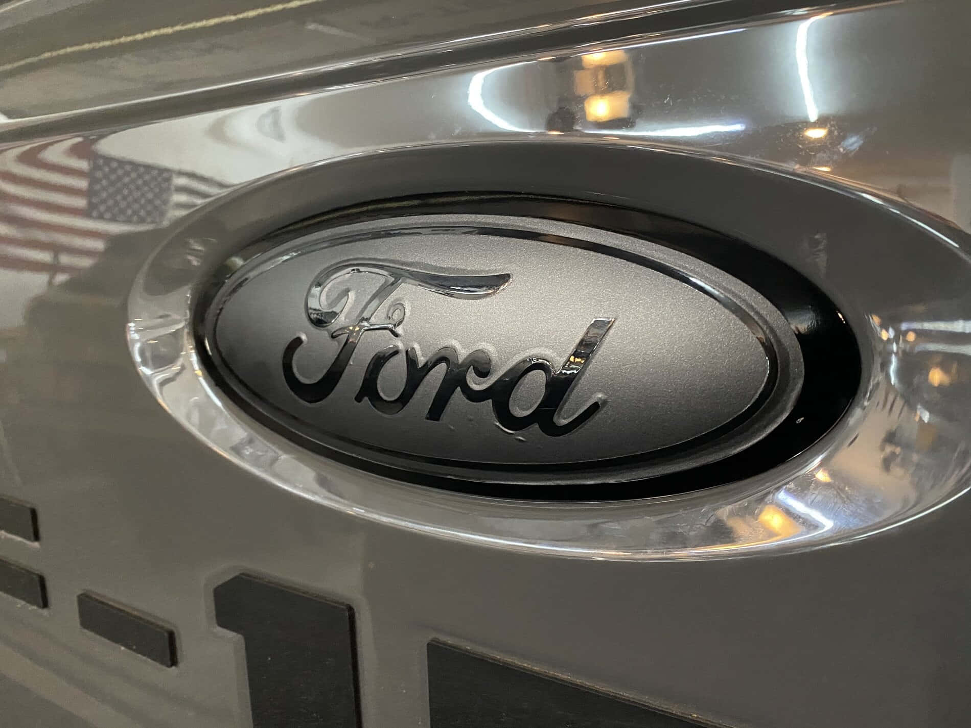 Ford Logo 桌布 壁紙 — 銀色引擎蓋上鍍鉻福特標誌的特寫，反射美國國旗和室內燈光。