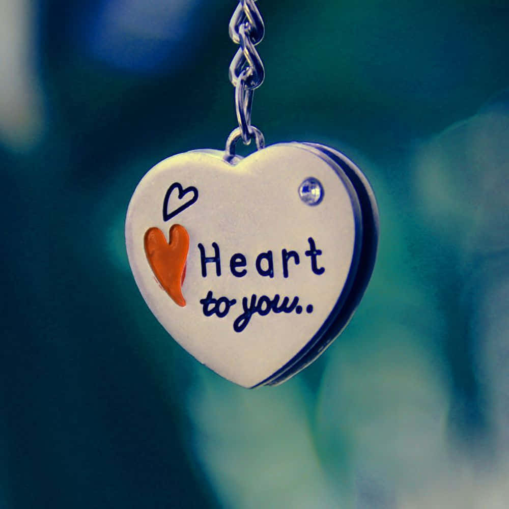 Whatsapp Dp 桌布 壁紙 — 銀色心形吊飾的特寫，上面刻有「Heart to you.」和一個橙色心形，掛在鏈條上，背景是模糊的藍綠色。