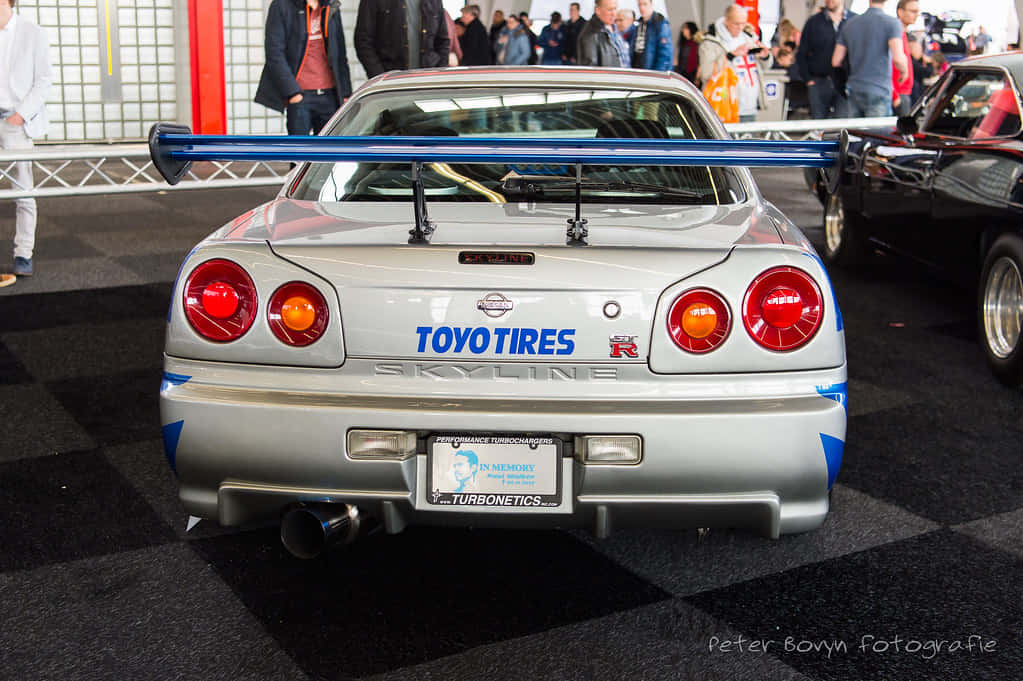 Paul Walker Skyline 桌布 壁紙 — 銀色日產Skyline R34，配備藍色尾翼和Toyo Tires塗裝，從後方觀看於車展中。